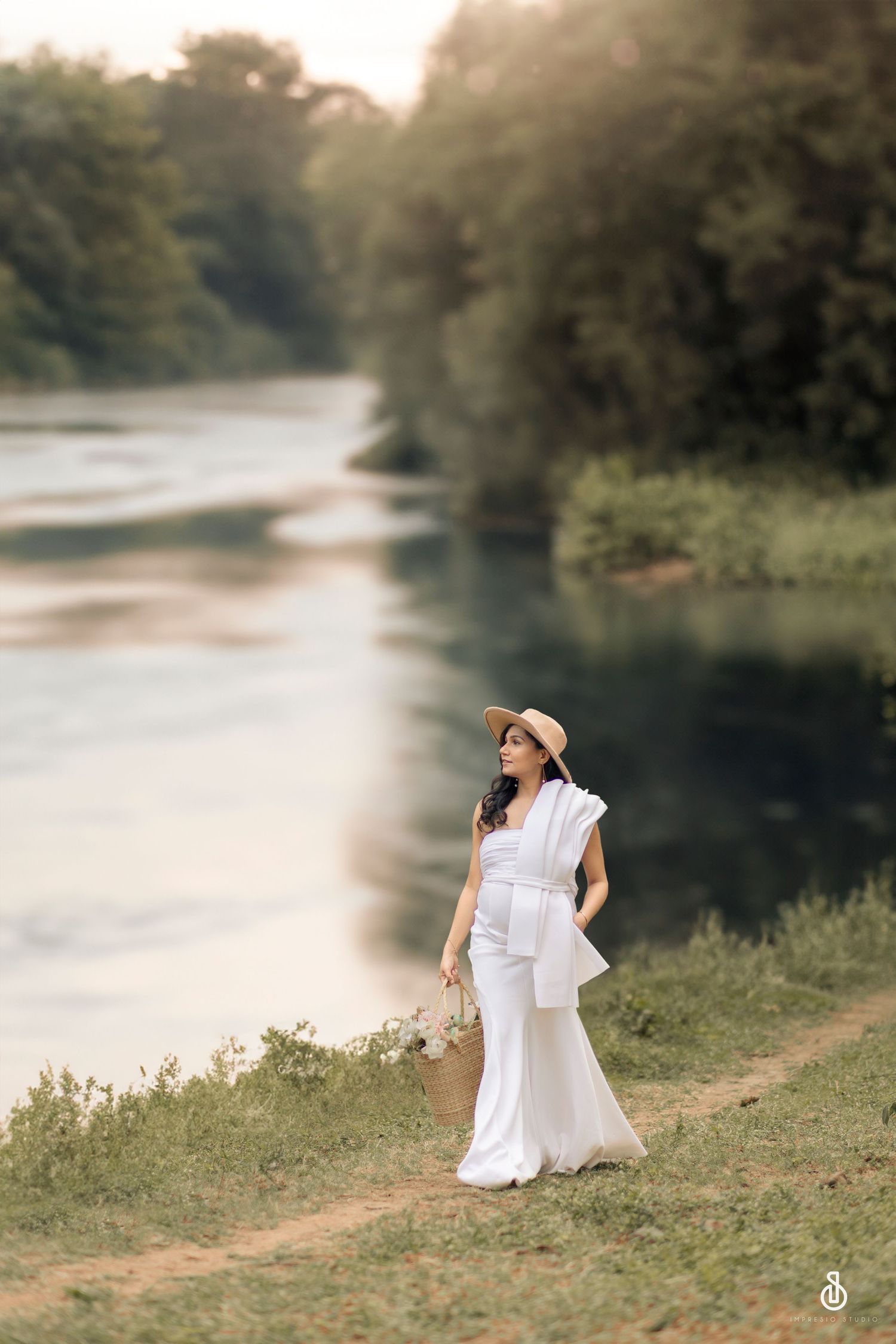 Woman in white outfit and straw hat walking along a scenic river path surrounded by lush green trees.