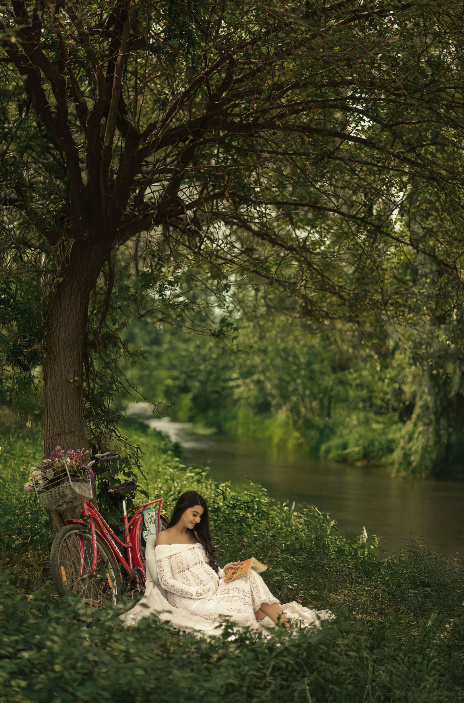 A peaceful riverside scene shows someone relaxing on a blanket next to a red bicycle under leafy tree canopy.