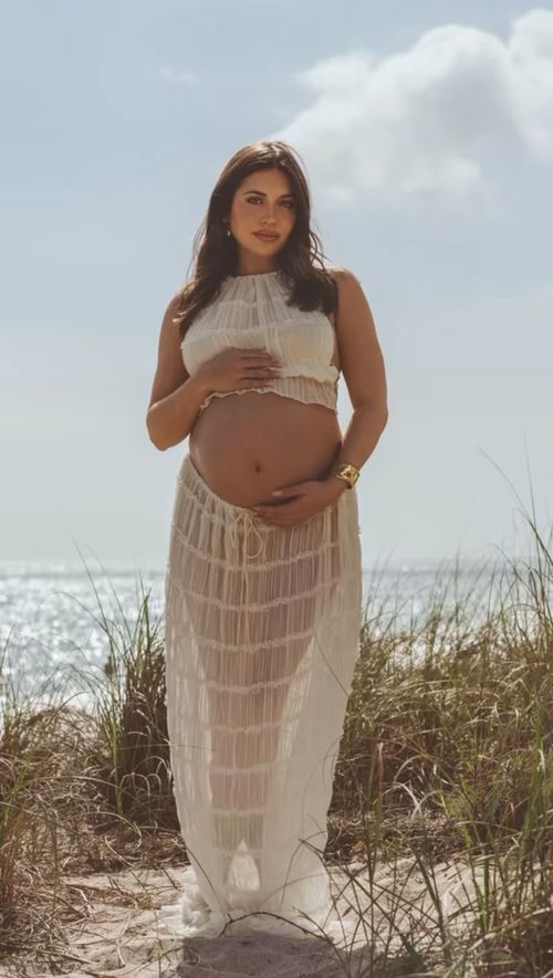 Expectant mother in white flowing dress poses for maternity photo on beach with ocean and sky in background.