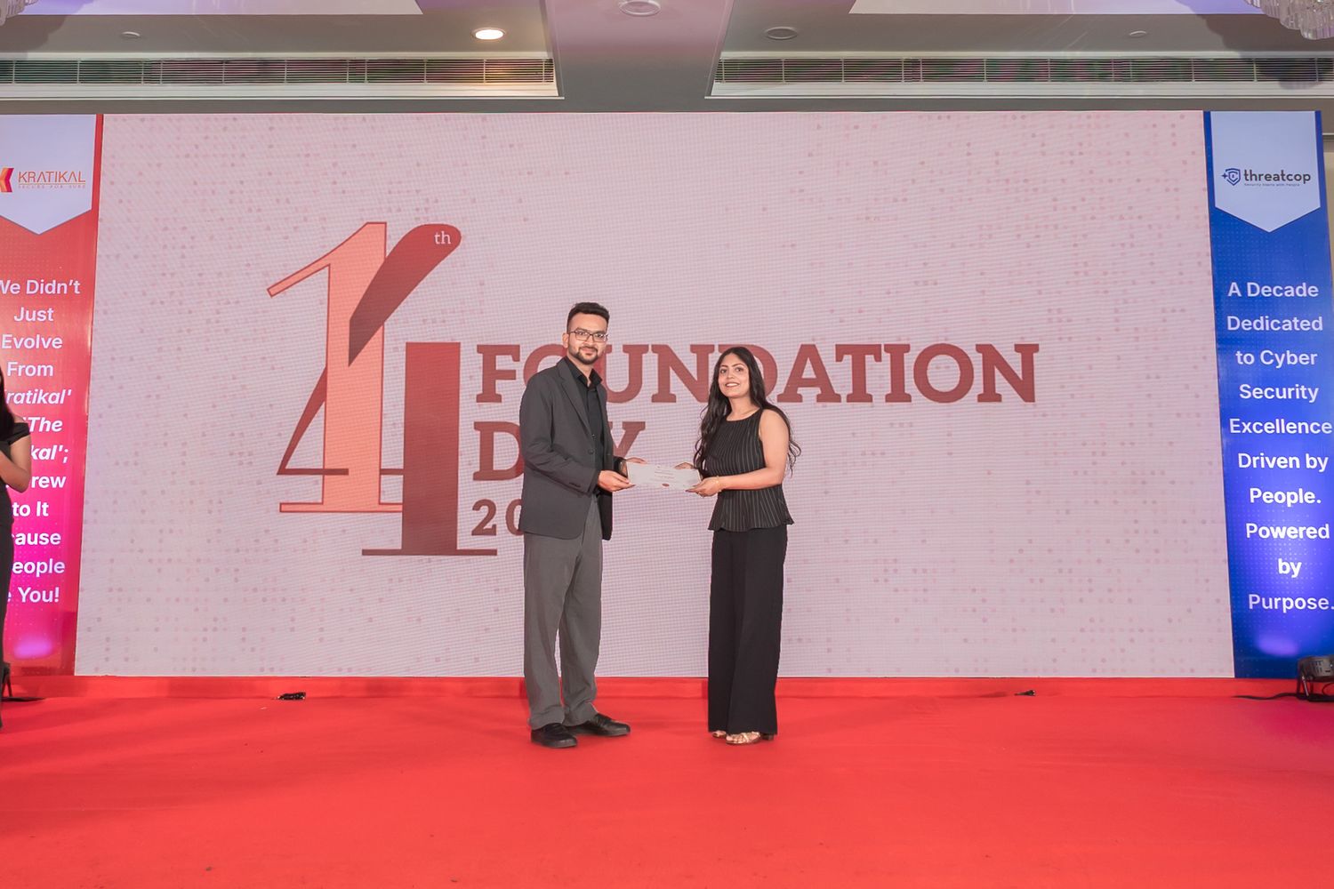 Man and woman in formal attire pose on a red carpet stage at a Foundation Day cybersecurity event, holding an award in front of a banner.