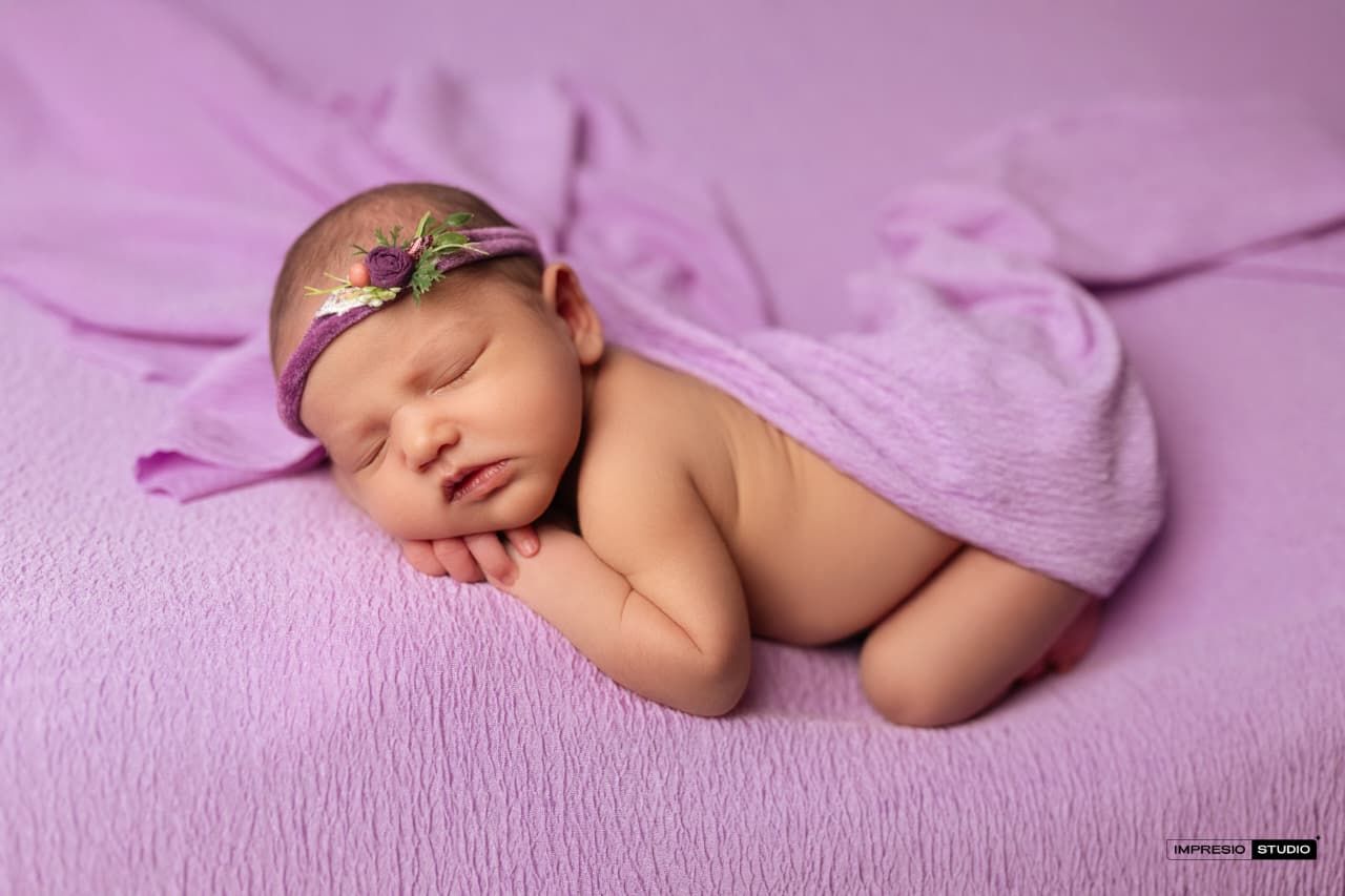 Newborn baby sleeping peacefully wrapped in purple blanket with floral headband on lavender background.