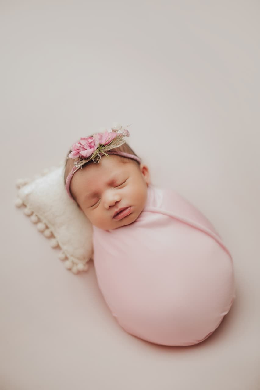 Sleeping newborn baby girl swaddled in pink wrap with floral headband resting on white pillow.