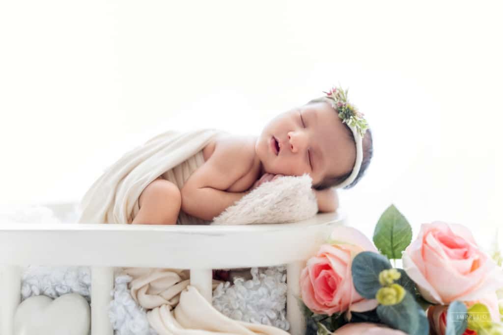 Newborn baby sleeping peacefully in white crib with floral crown and pink roses nearby.