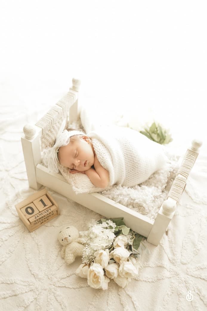 Sleeping newborn baby wrapped in white knit swaddle on miniature bed with teddy bear and white roses.
