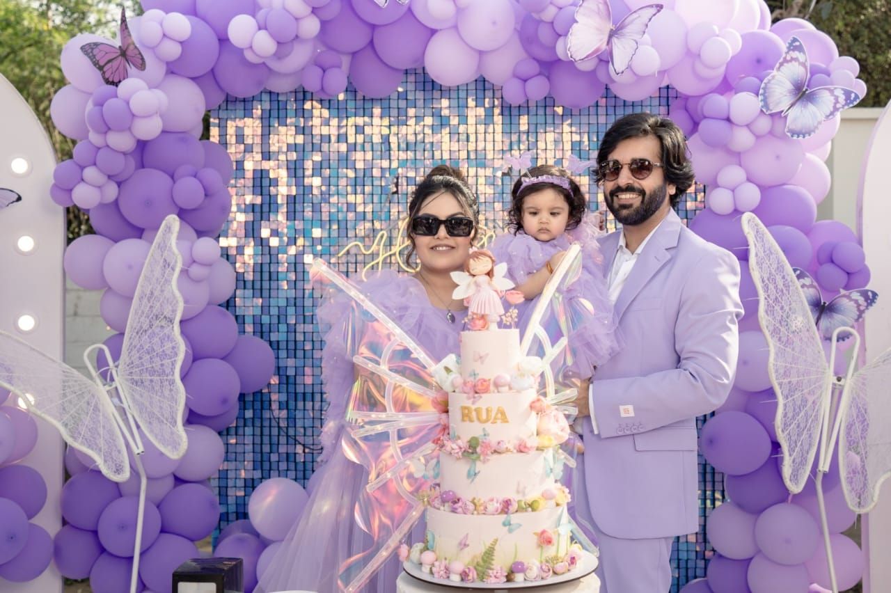 Family celebrating a first birthday with a butterfly-themed cake under a purple balloon arch.