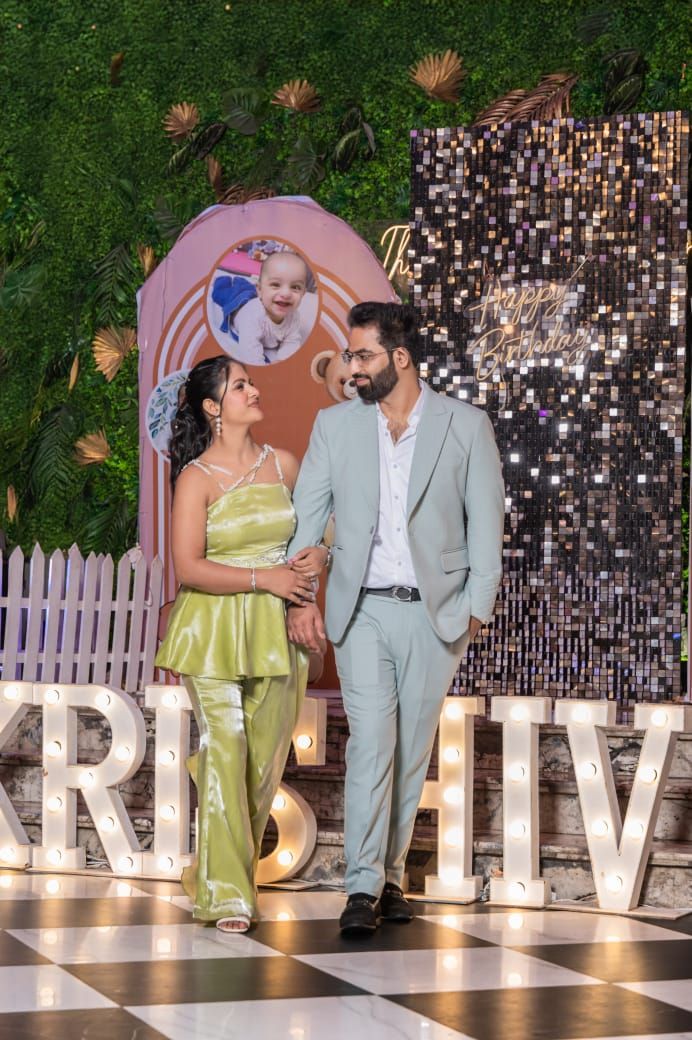Couple in elegant attire posing at a festive Krishna-themed birthday party backdrop with marquee letters.