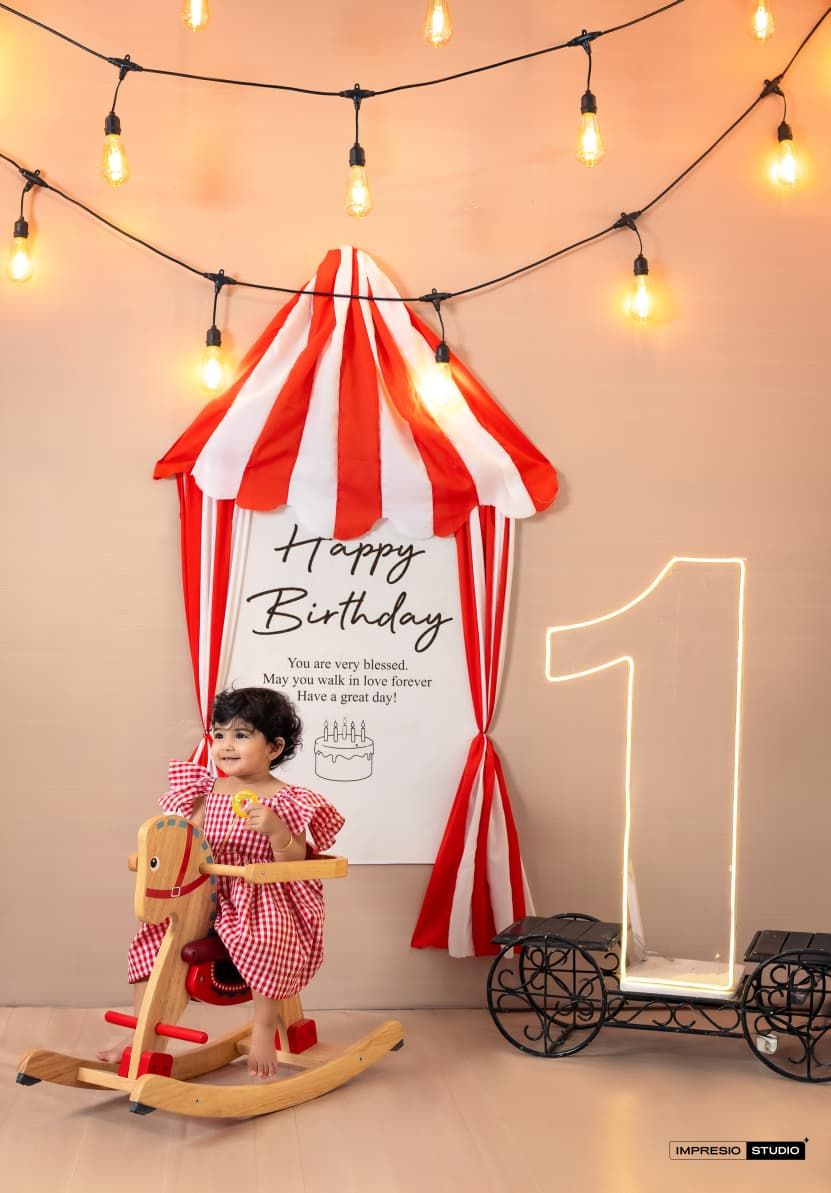Toddler girl in red gingham dress sits on rocking horse at circus-themed first birthday photo setup.