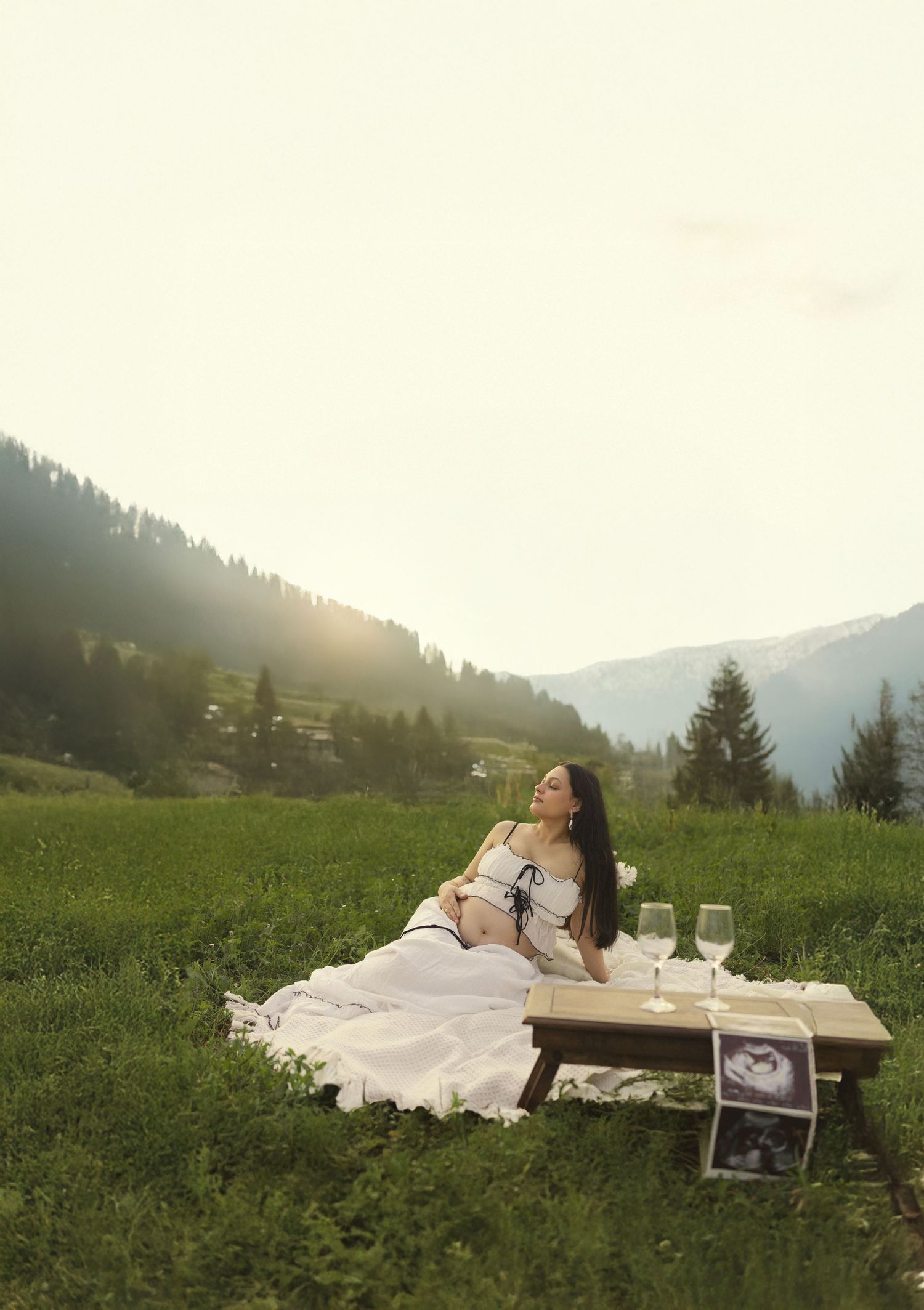 Pregnant woman relaxing on a blanket in a green alpine meadow at sunset with wine glasses and ultrasound photo.