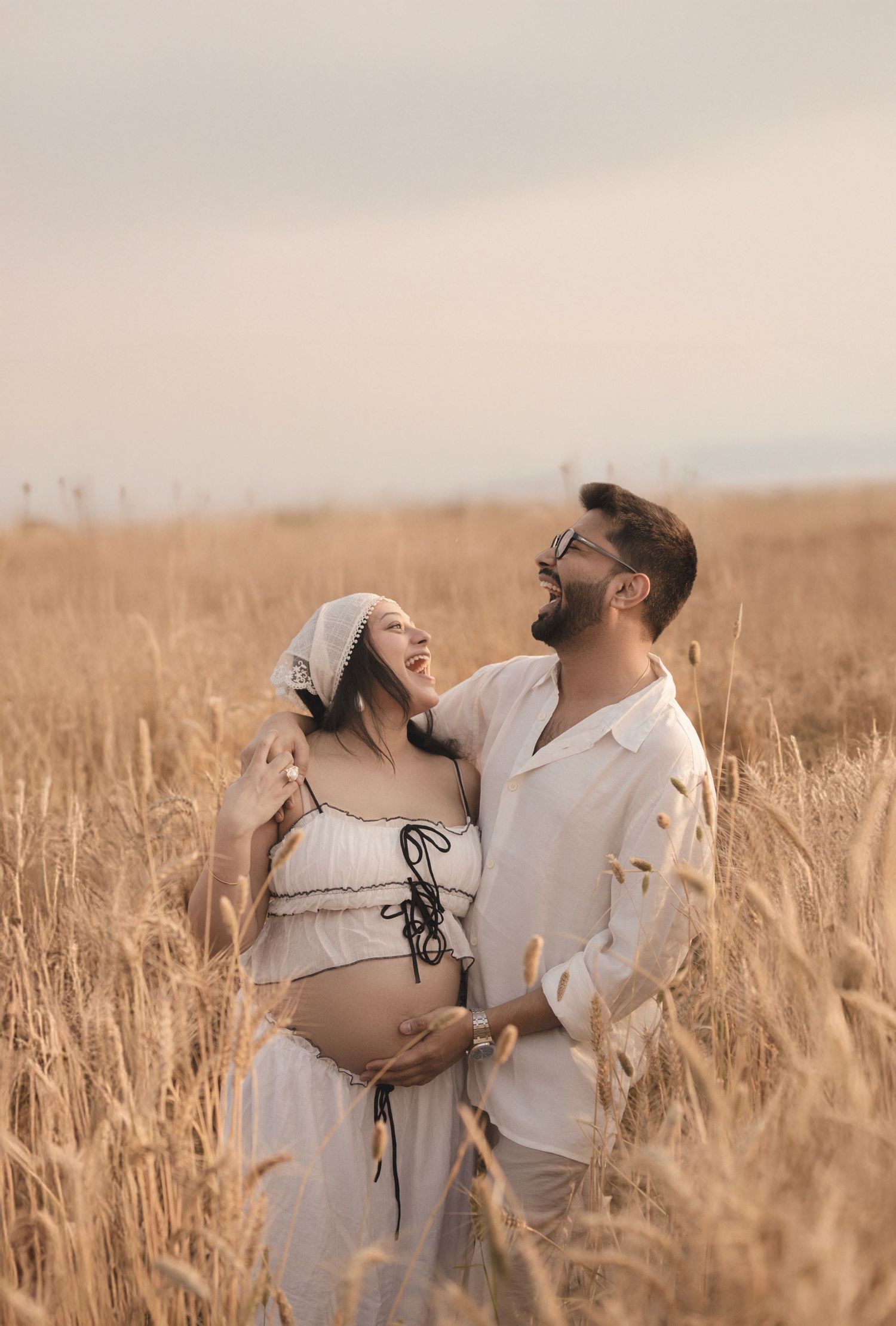 Couple in white outfits embracing in golden wheat field, pregnant woman cradling baby bump, maternity photo shoot.