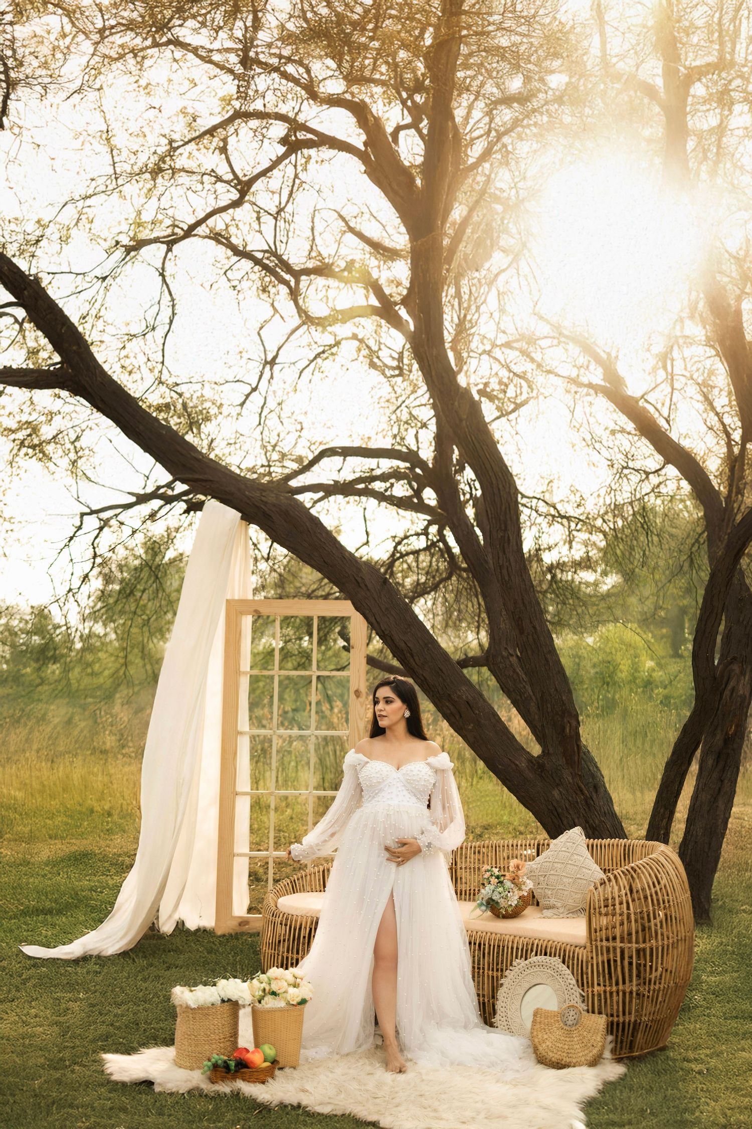 Pregnant woman in white lace gown poses outdoors near rustic decor and large tree in golden sunset light.