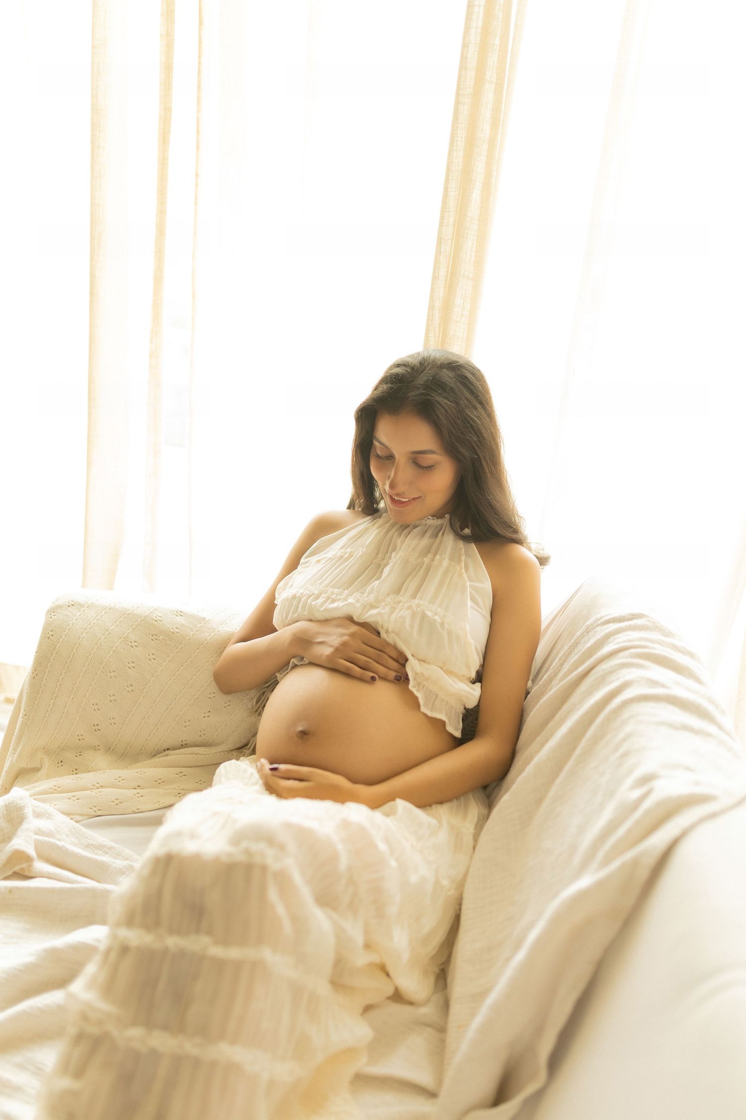 Expectant mother in white dress rests peacefully on white bedding while cradling her pregnant belly in soft window light.