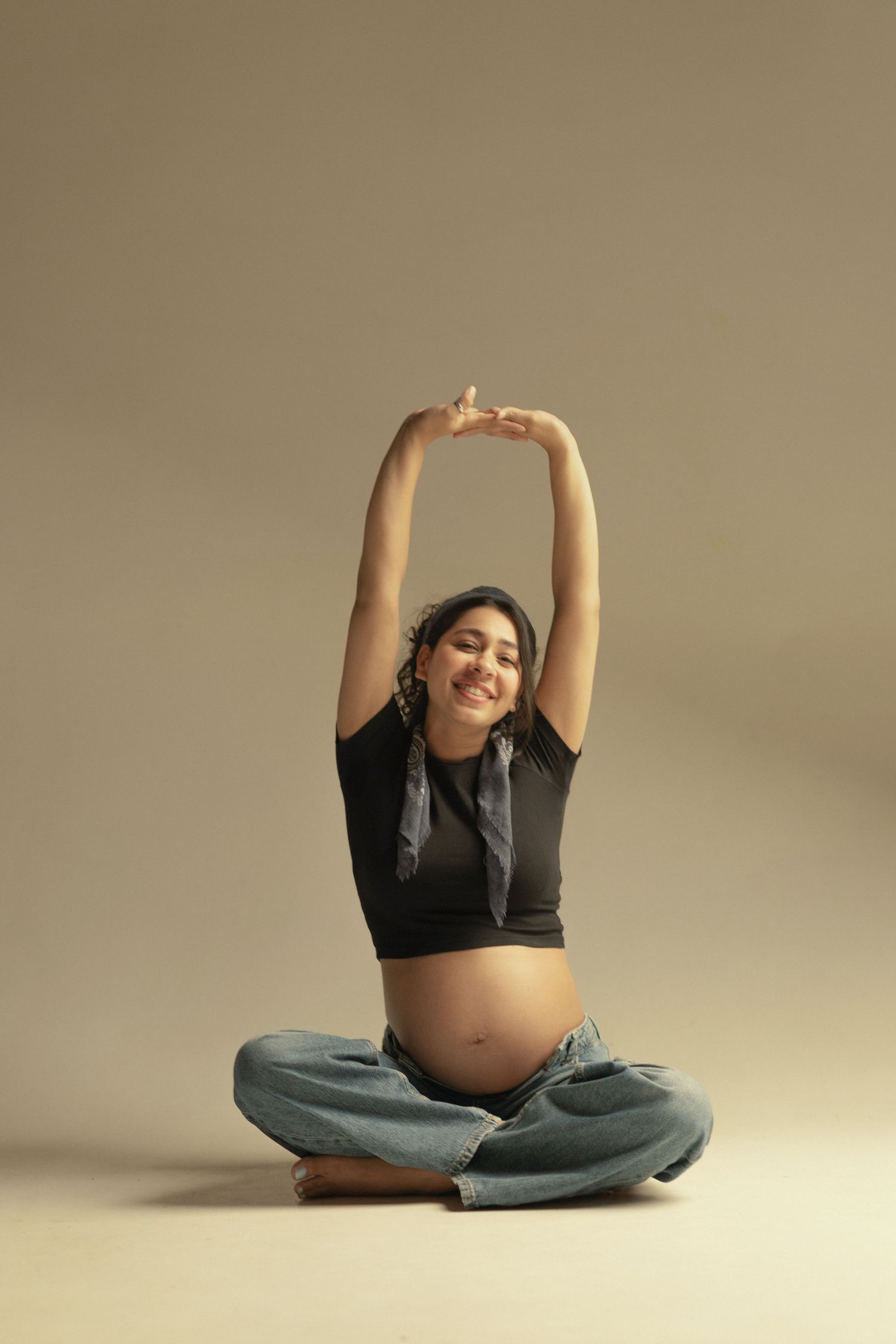 A pregnant person in a black crop top and jeans sits cross-legged on the floor while stretching their arms overhead.