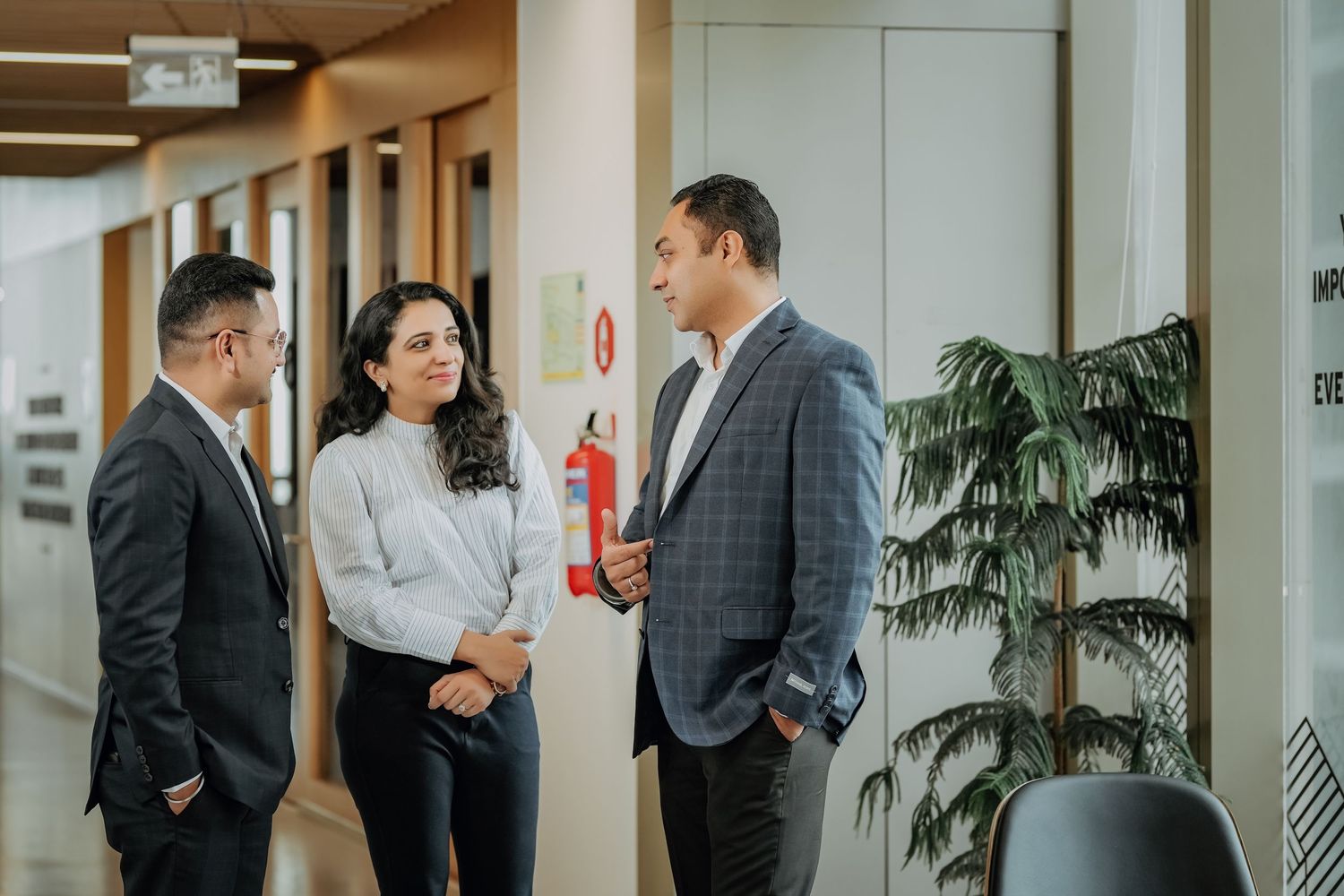 Business professionals gather in a modern office hallway for informal discussions near indoor plants and windows.