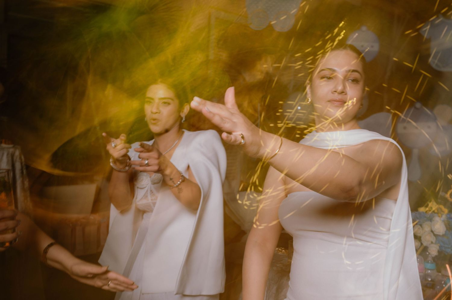 Two women in white outfits celebrate at a festive event with golden confetti and warm lighting.