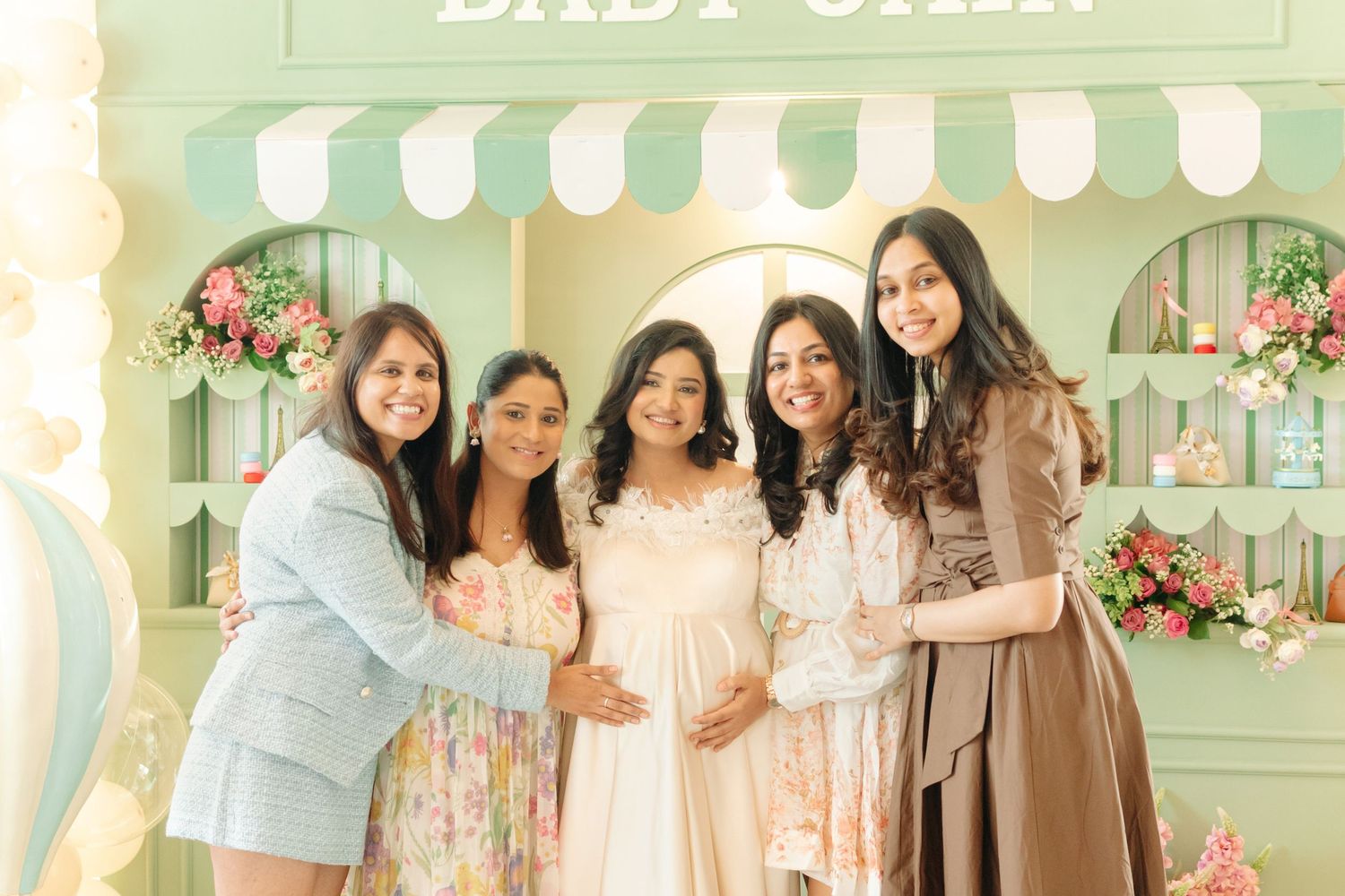 Five women pose together at a pastel green baby shower backdrop with floral decorations and a pregnant woman in white.