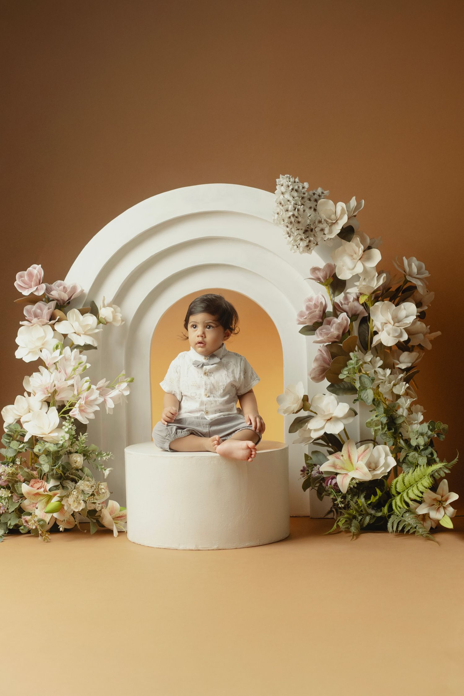 Beautiful floral arch setup with white flowers and greenery surrounding a round pedestal against a warm brown backdrop.