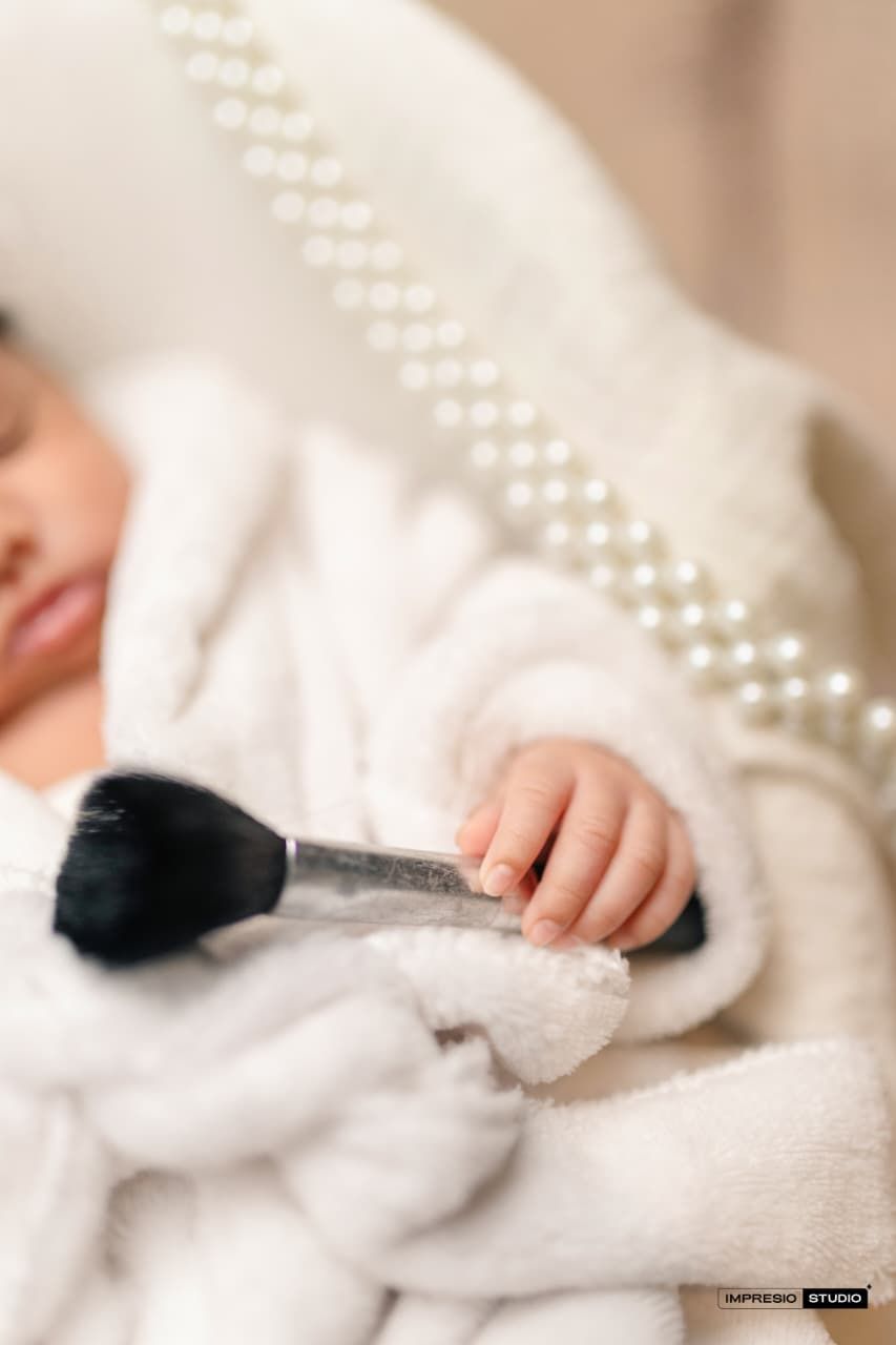 Newborn baby in white robe holding a makeup brush, surrounded by pearl necklace, soft focus photography.