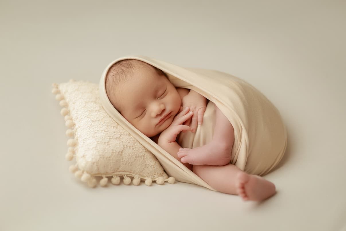 Peaceful newborn baby sleeping wrapped in a cream blanket on a small pillow in a soft studio setting.