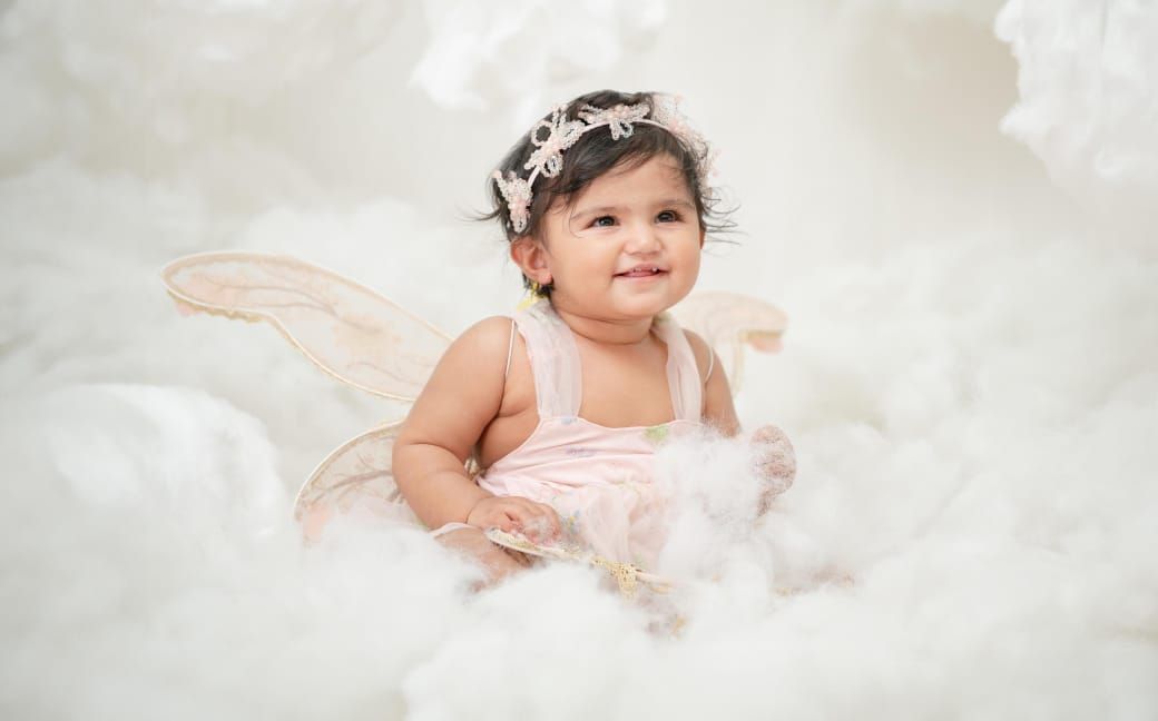 Adorable baby girl in pink dress with fairy wings and floral headband smiling among fluffy white clouds.