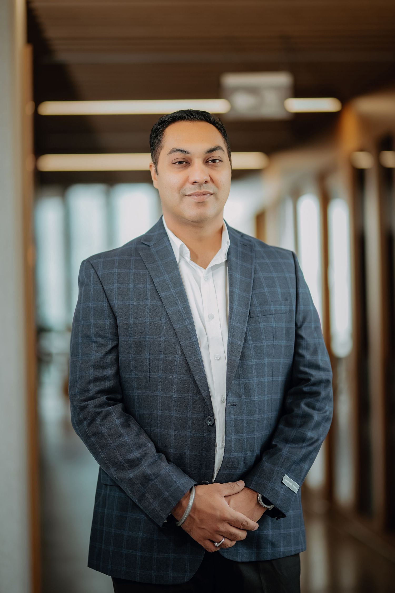 Professional wearing a navy windowpane suit and white shirt poses in an office hallway with blurred background.