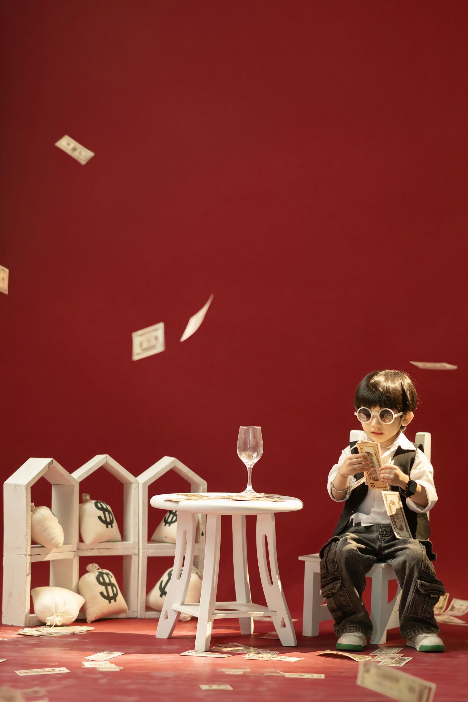 A child in sunglasses sits counting money with bills falling around them near white shelves with money bags on red background.