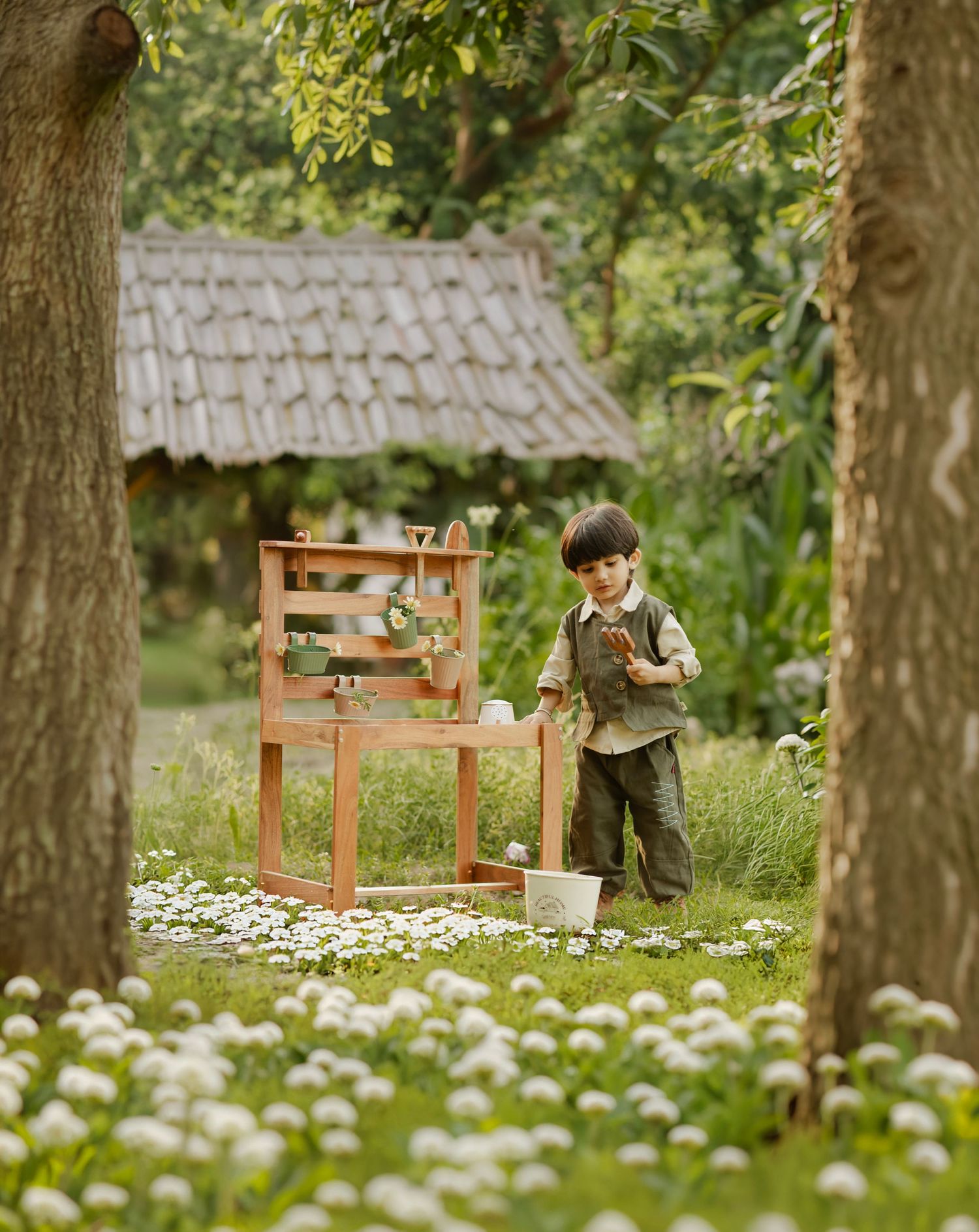 Young boy playing in a lush garden beside a wooden shelf, surrounded by white flowers and a rustic thatched cottage.