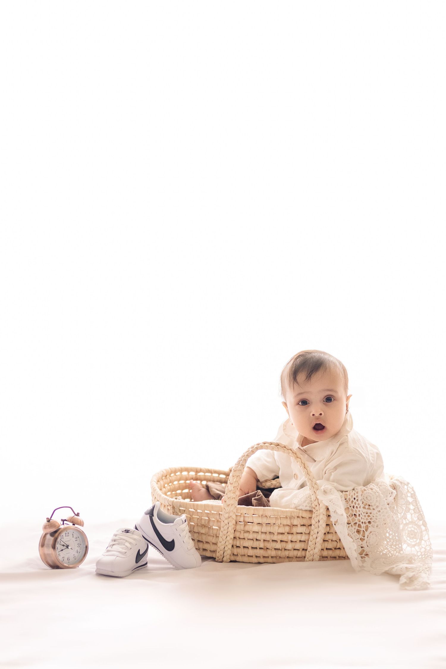 Baby in white dress sits in woven basket next to Nike sneakers and pink alarm clock on white background.