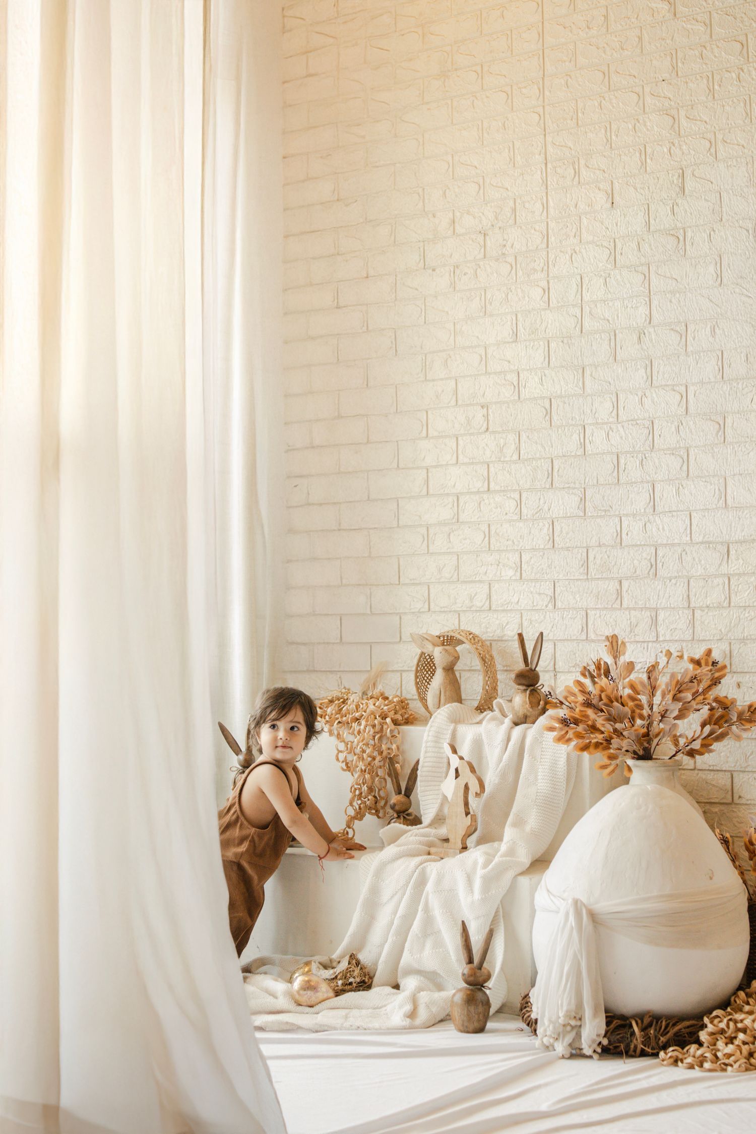 Young child in brown romper plays among Easter bunny decorations and dried flowers in a bright boho-style white room.