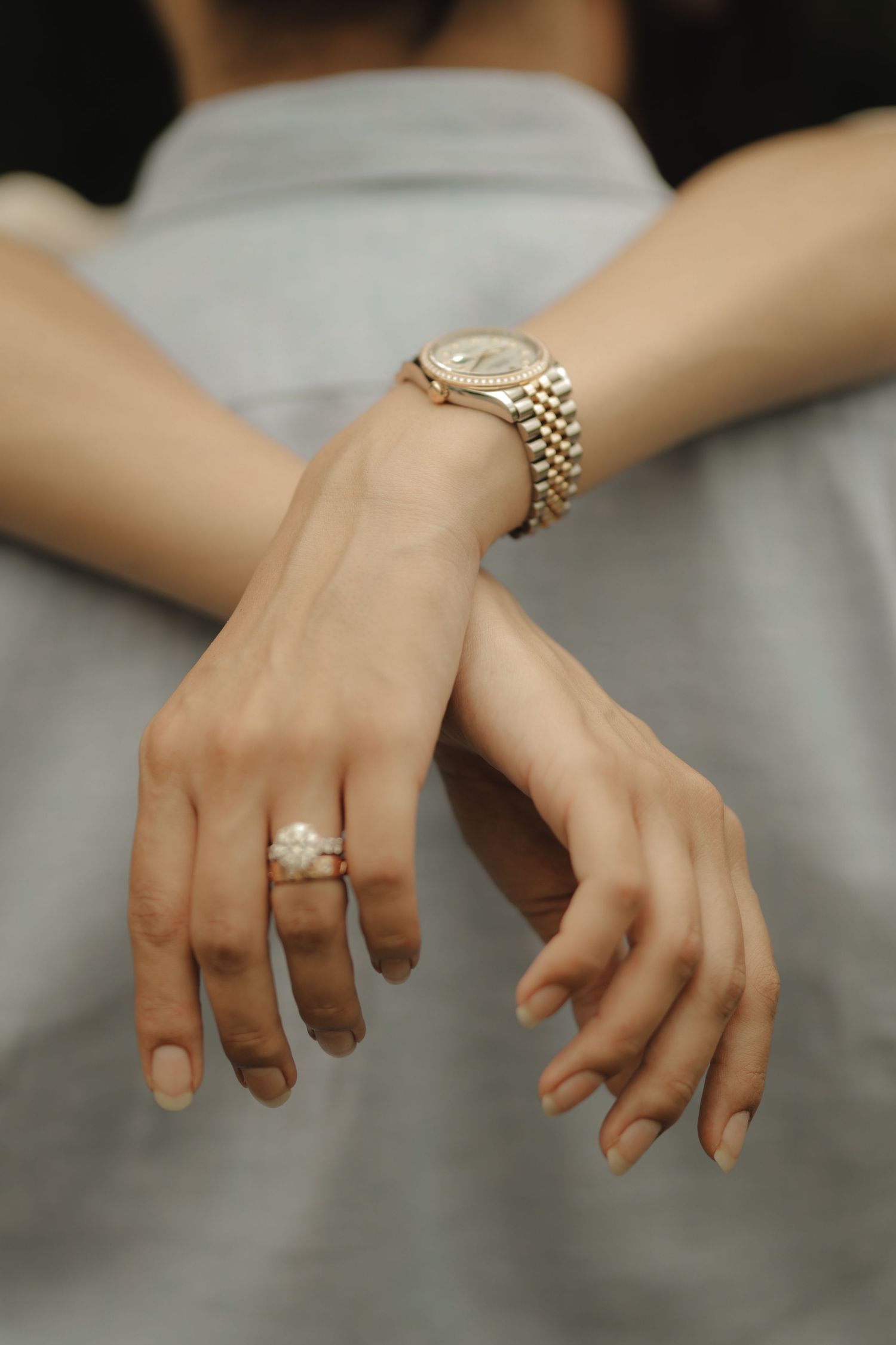 Close-up of crossed hands wearing a gold Rolex watch and floral diamond engagement ring on a light blue dress.