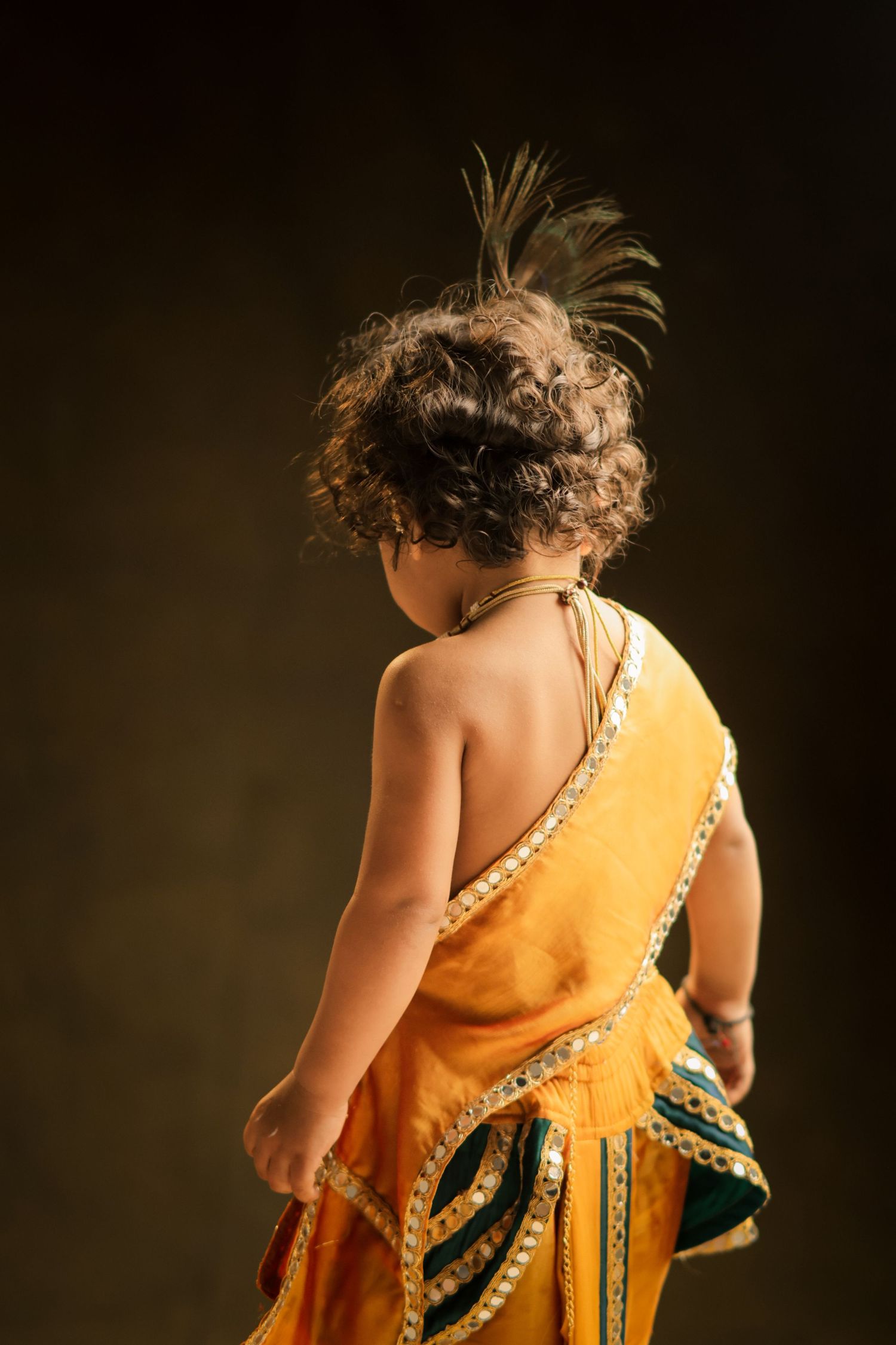 Young child dressed in traditional Indian attire with peacock feather hair accessory, viewed from behind.