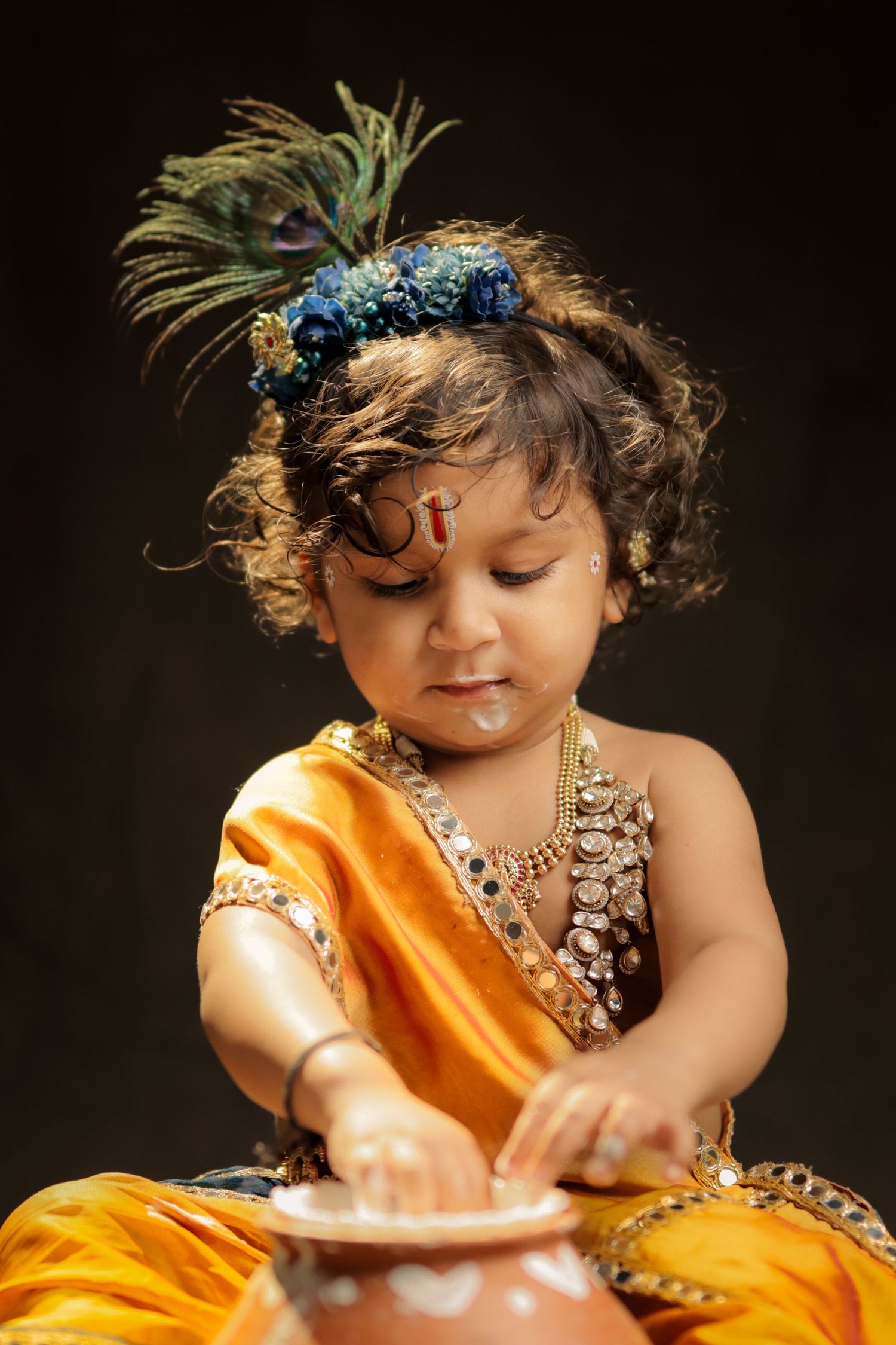 Baby dressed as Lord Krishna in yellow silk, peacock feather crown, gold jewelry, reaching into clay pot.