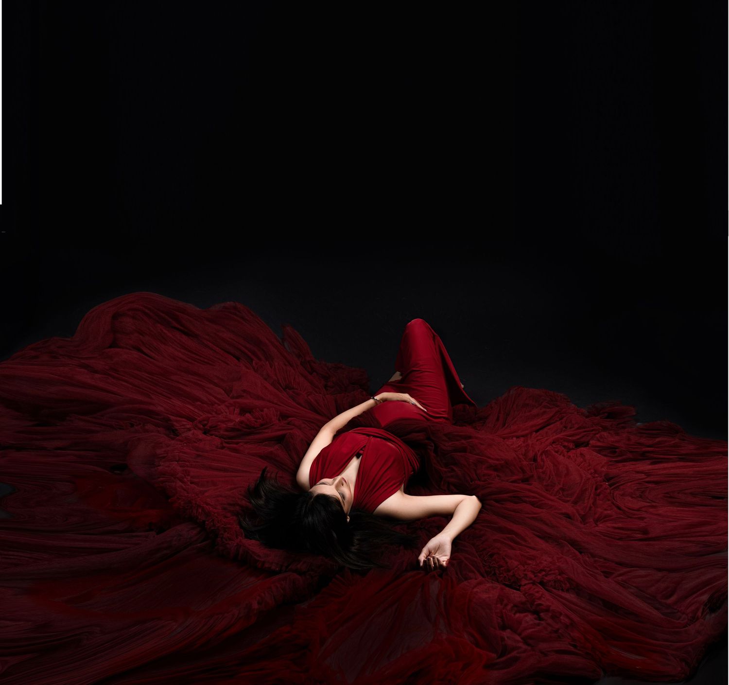A dramatic photo of a figure in a flowing red dress lying on dark fabric against a black background.