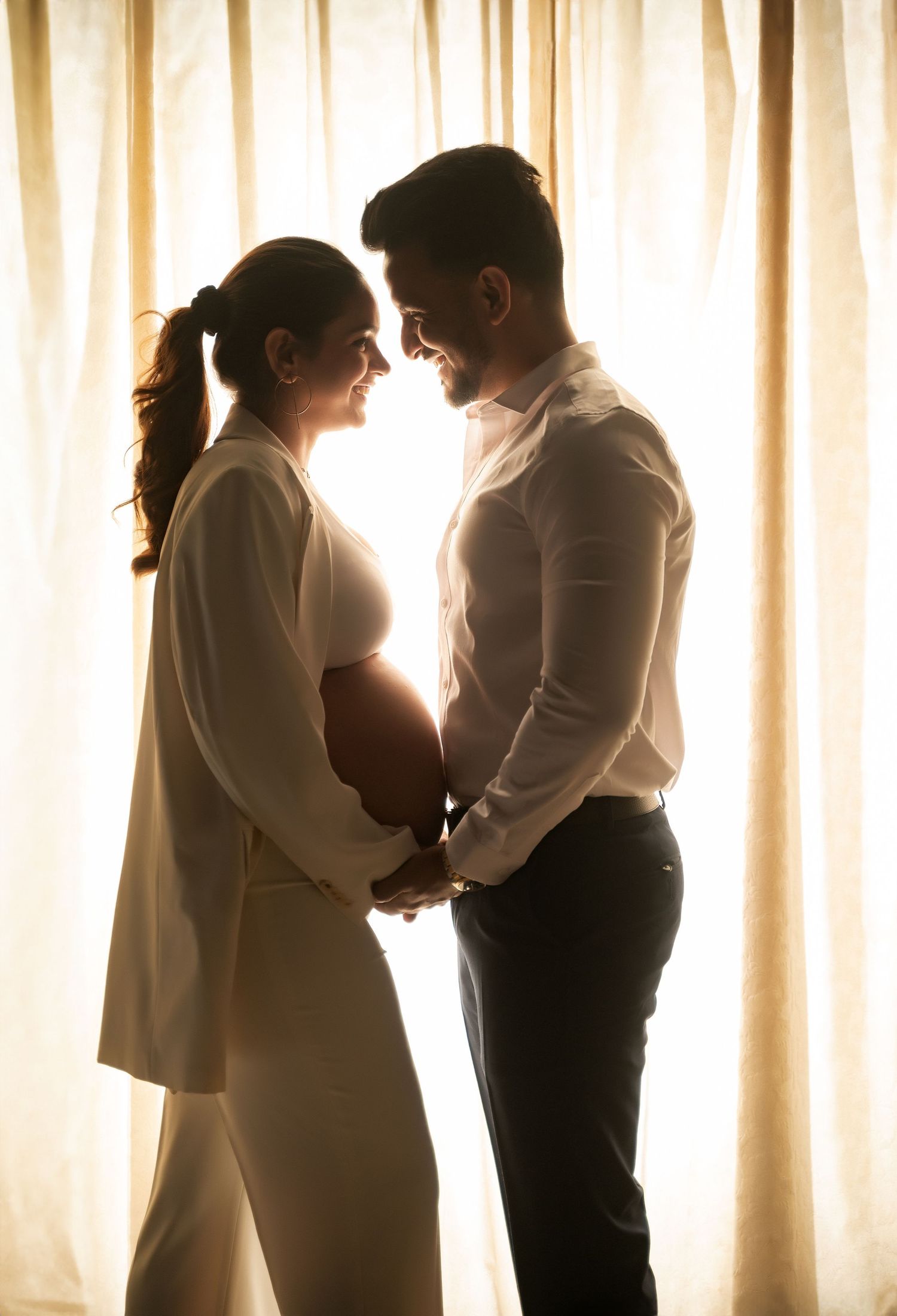 Silhouetted couple sharing intimate moment in front of sunlit curtains during maternity photoshoot.