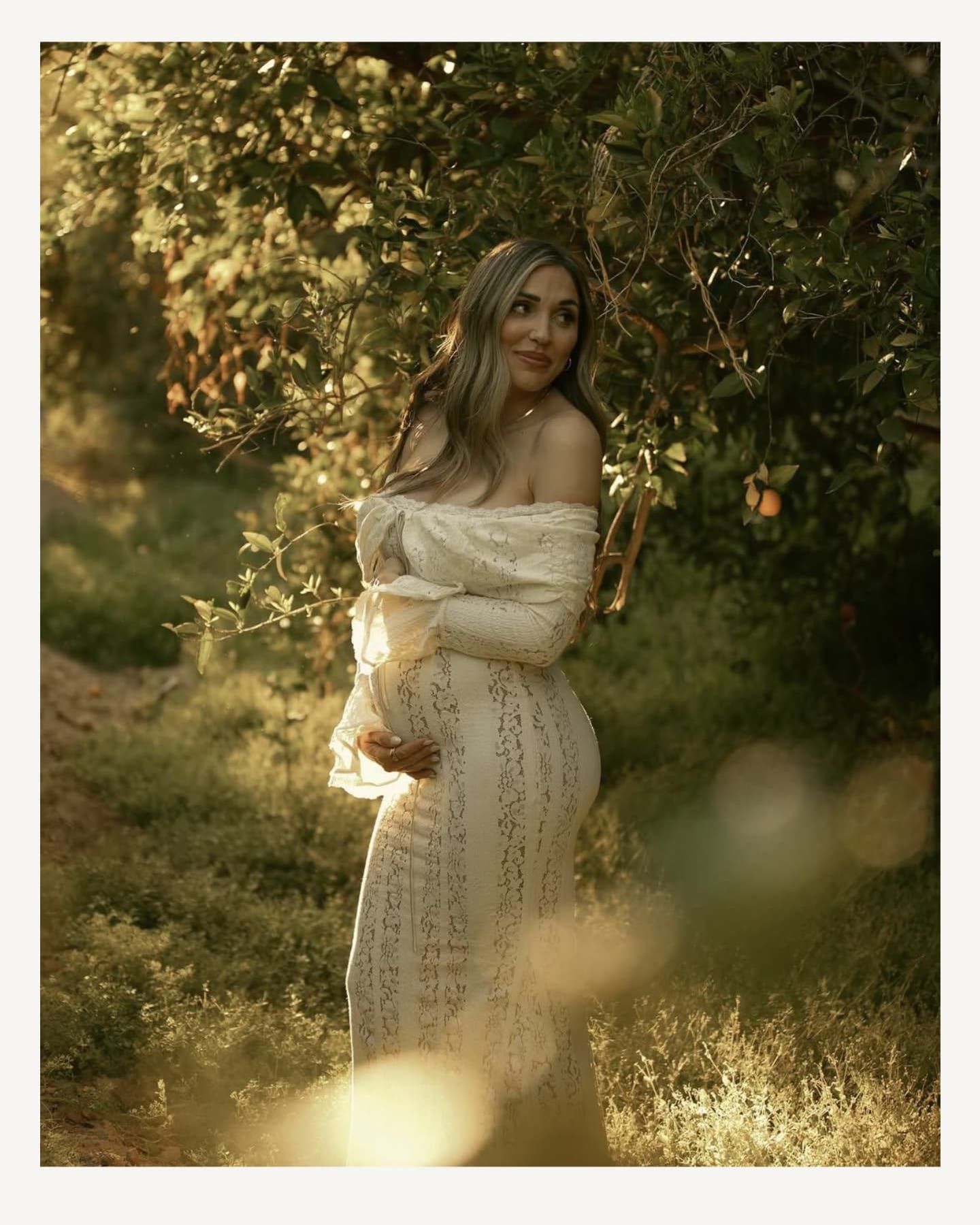 Romantic maternity photoshoot in a sunlit orchard with flowing white lace dress.