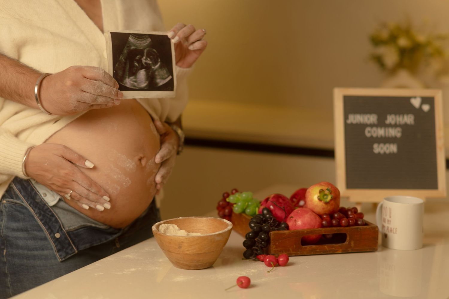 Pregnant woman holding ultrasound photo near fruit display and Junior Johnni Coming Soon sign.