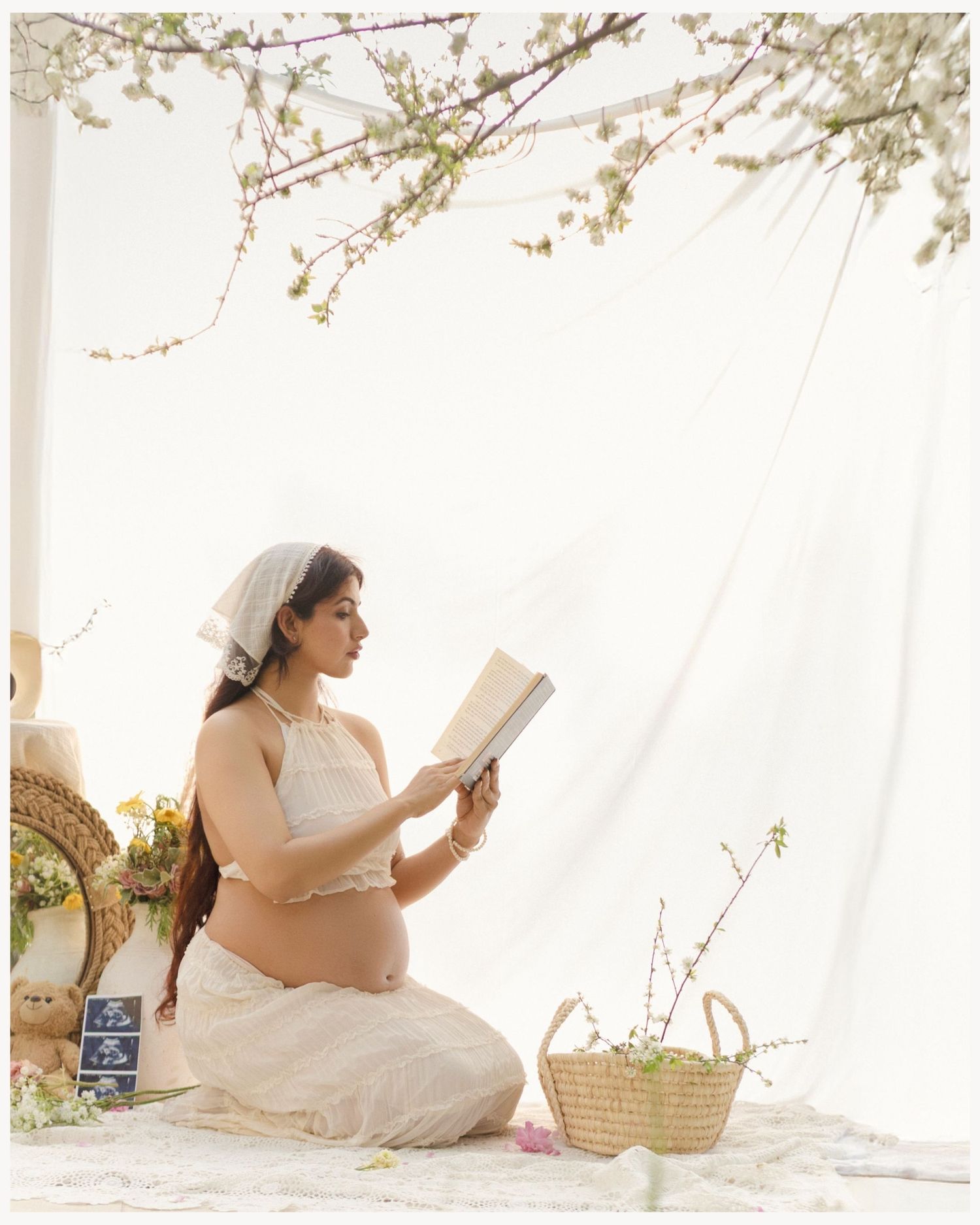 Pregnant woman in white crop top and skirt reads a book outdoors, surrounded by flowers and a wicker basket.