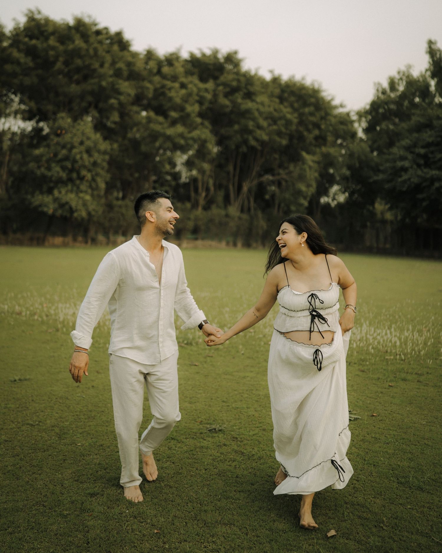 Couple in matching white outfits walking hand in hand barefoot on green grass with trees in background.