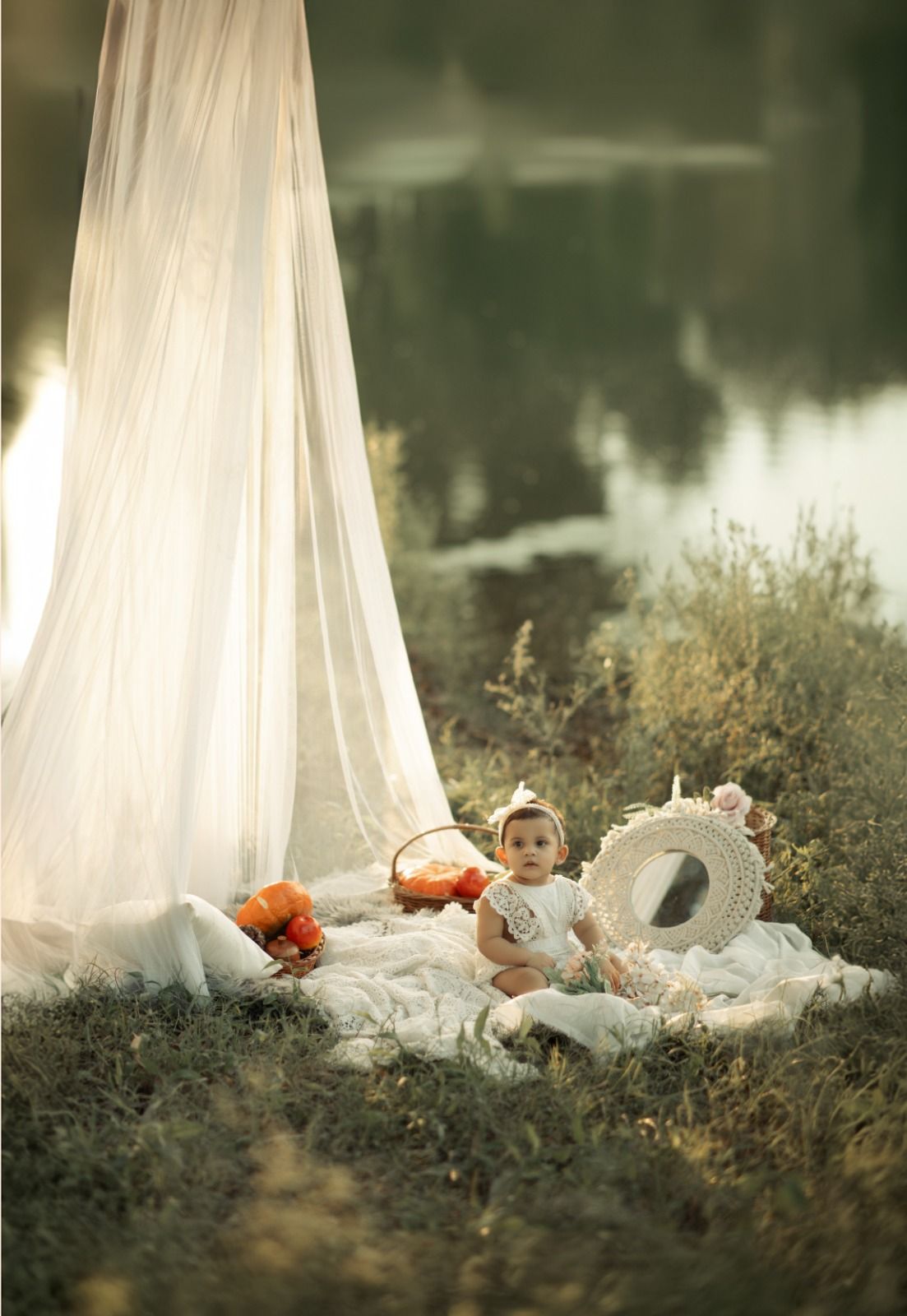 Baby sitting on white blanket outdoors near sheer canopy with hat and basket by a serene lake.
