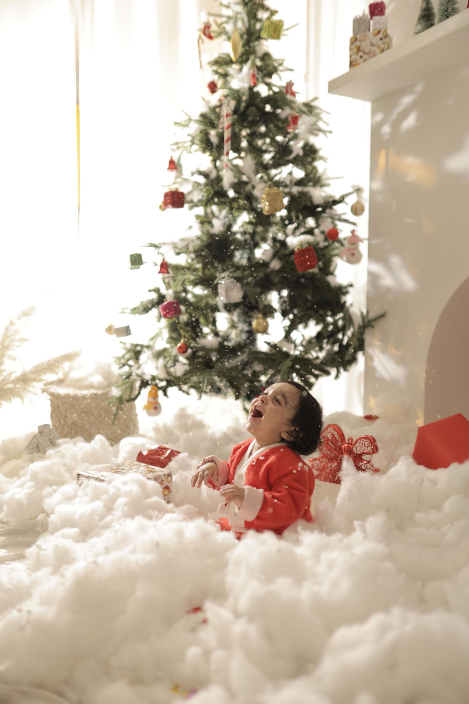 Child in red pajamas laughing while lying in artificial snow near a decorated Christmas tree indoors.