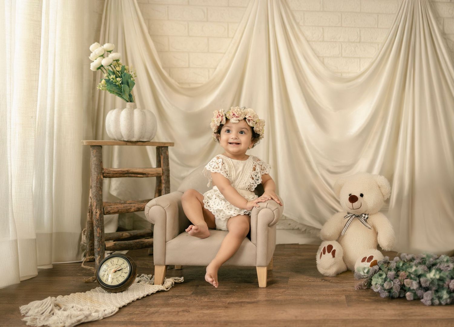 Baby girl in floral headband sits on mini sofa with teddy bear, clock, and white drape backdrop.