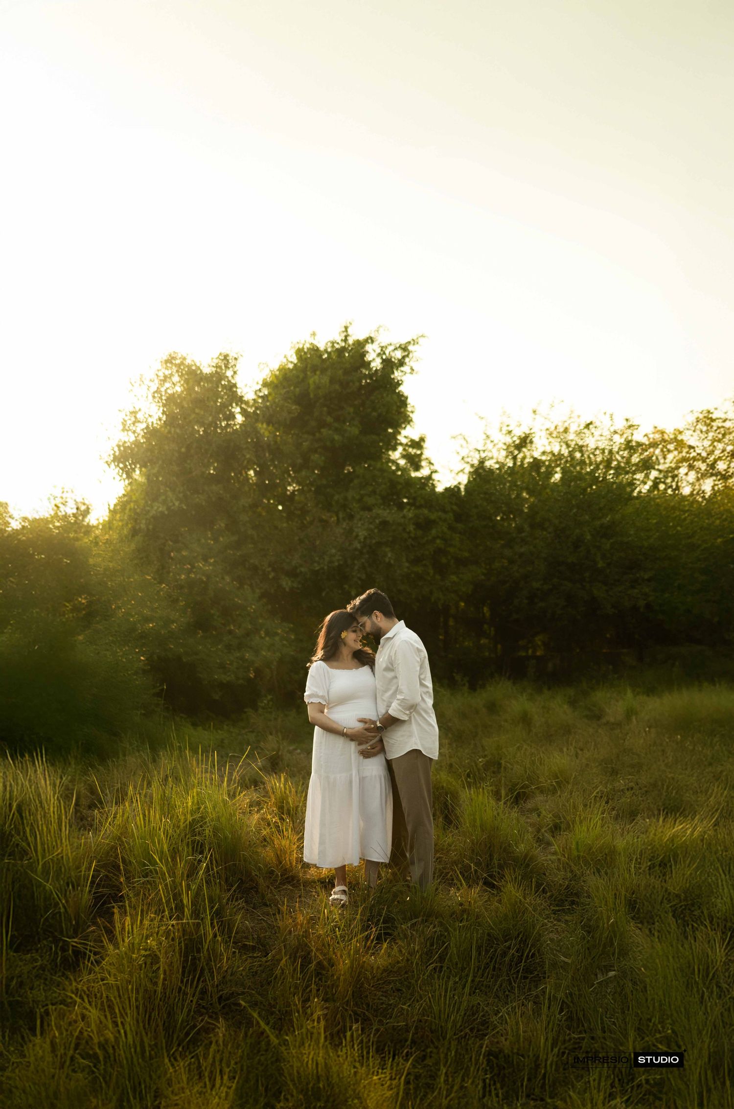 Couple embracing in a sunlit green meadow with trees in the background during golden hour.