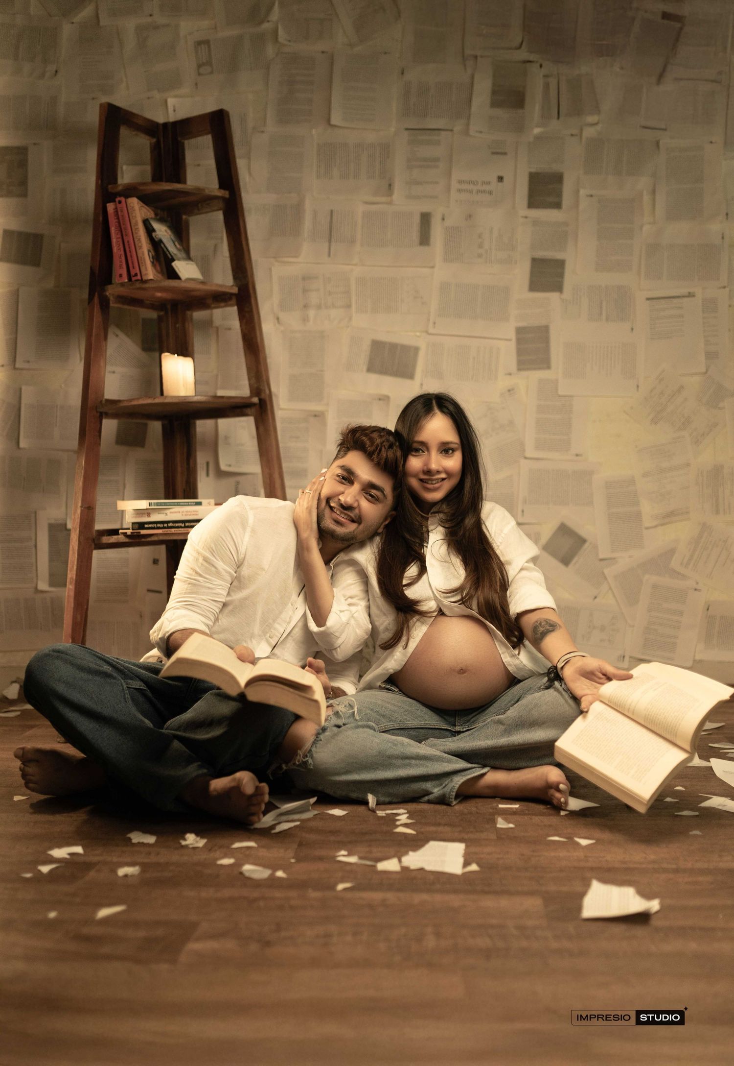 Couple sitting on floor with open books, pregnant woman smiling, ladder bookshelf, pages on wall backdrop.