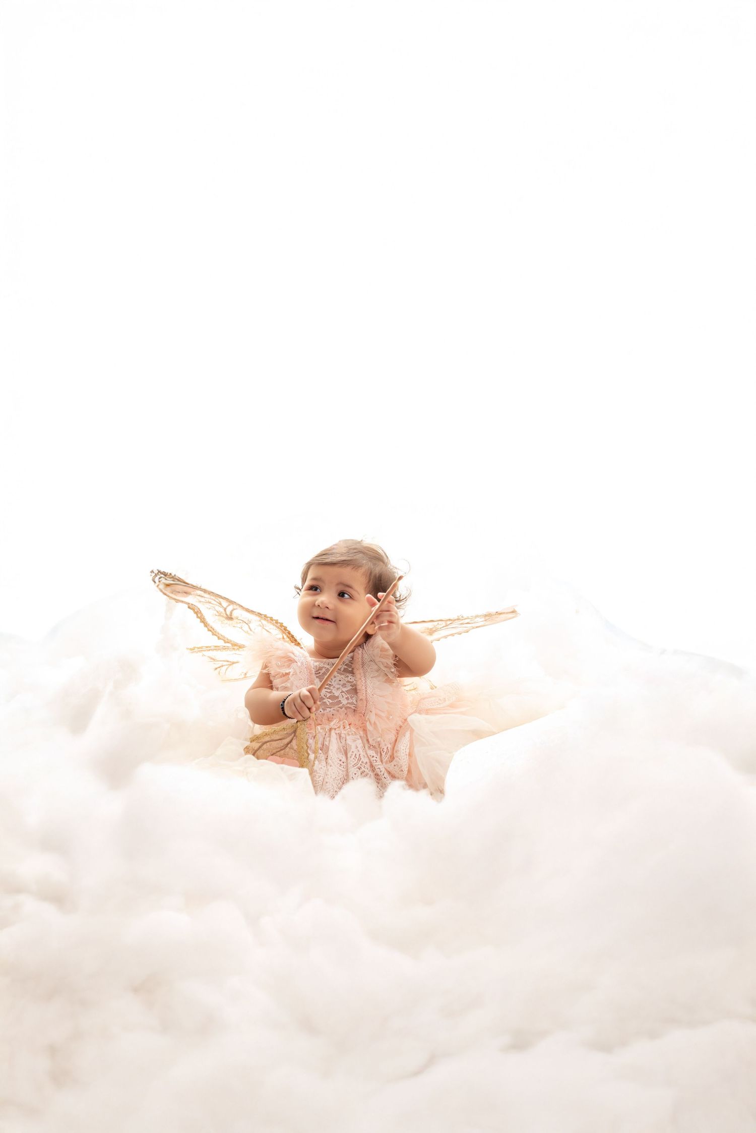 Baby girl in pink dress with gold fairy wings sits among fluffy white clouds in a dreamy portrait photo.
