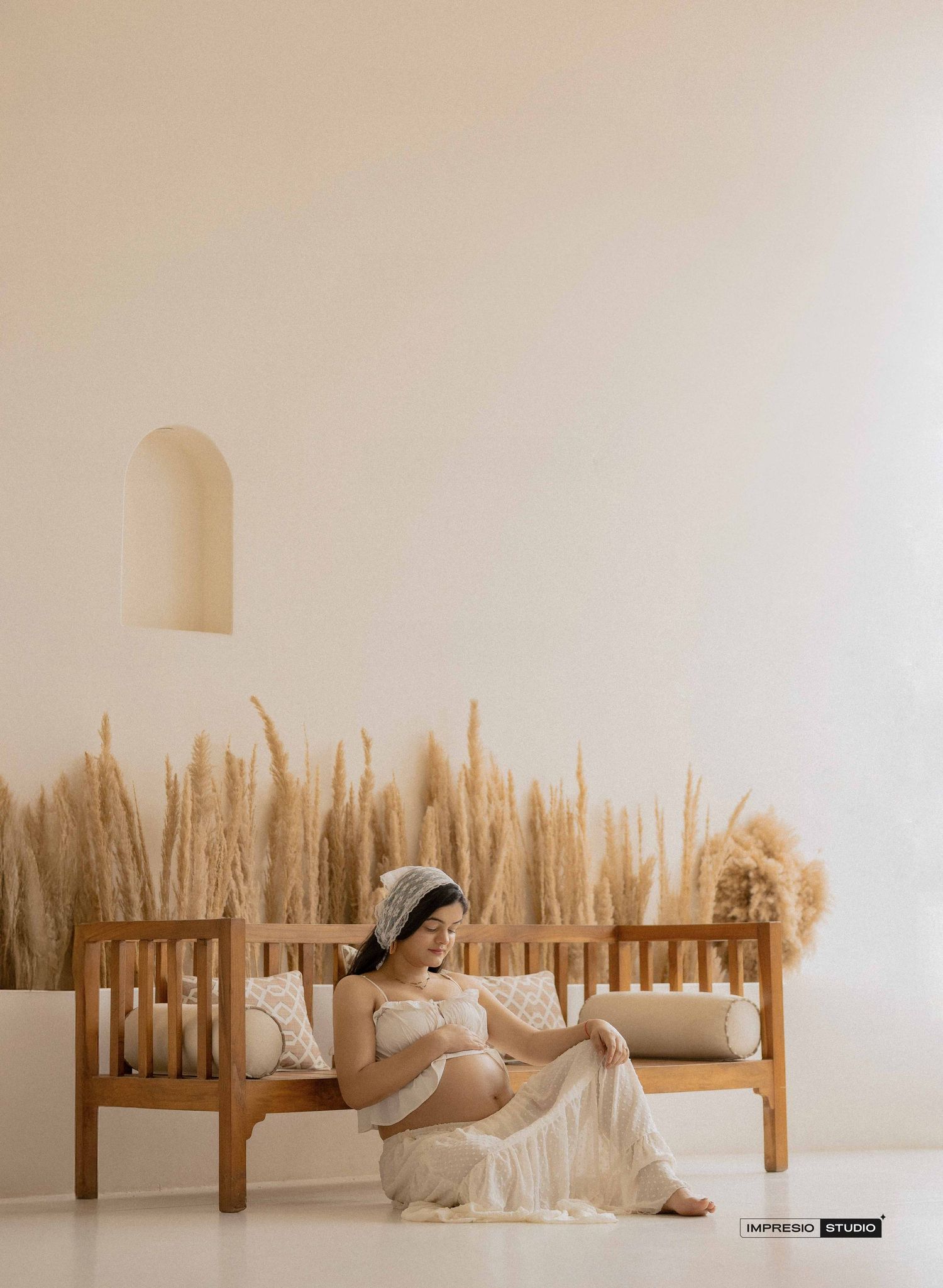 Pregnant woman in white lace dress sits on wooden bench with pampas grass backdrop in minimalist studio.