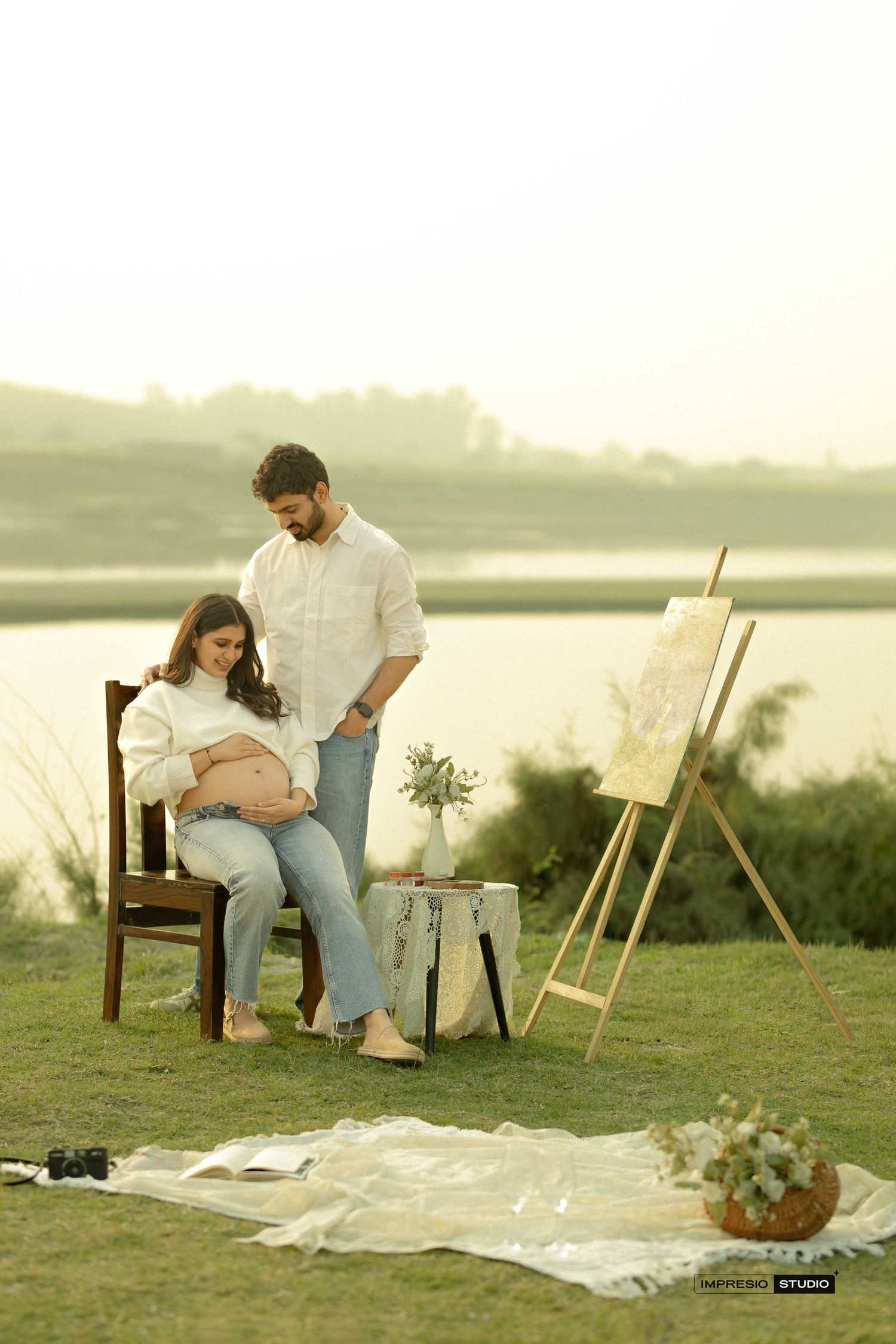 Couple poses outdoors for maternity photoshoot near lake, woman seated showing baby bump, easel and flowers nearby.