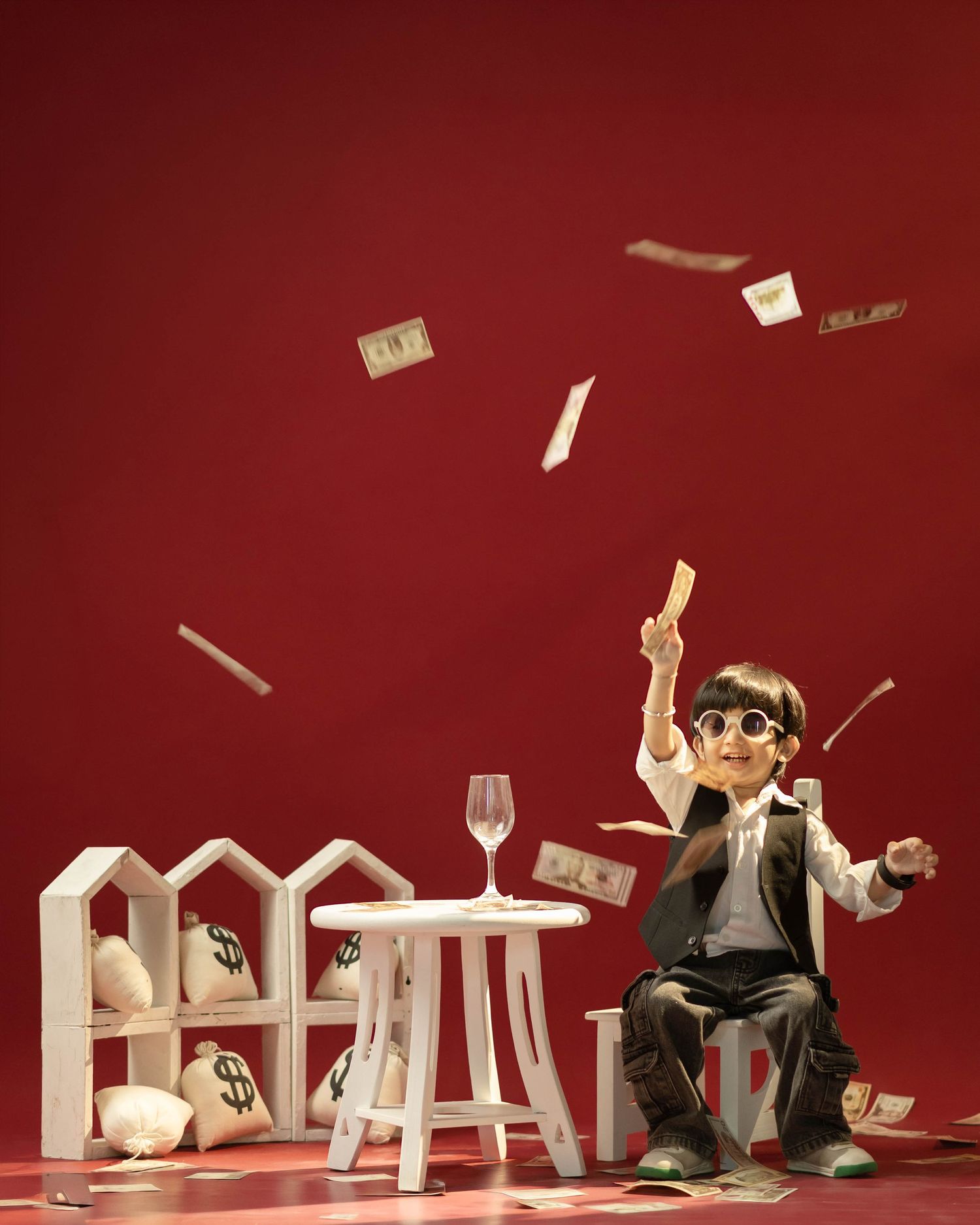 A small figurine in a suit and sunglasses sits throwing money in the air near white miniature houses and a wine glass.