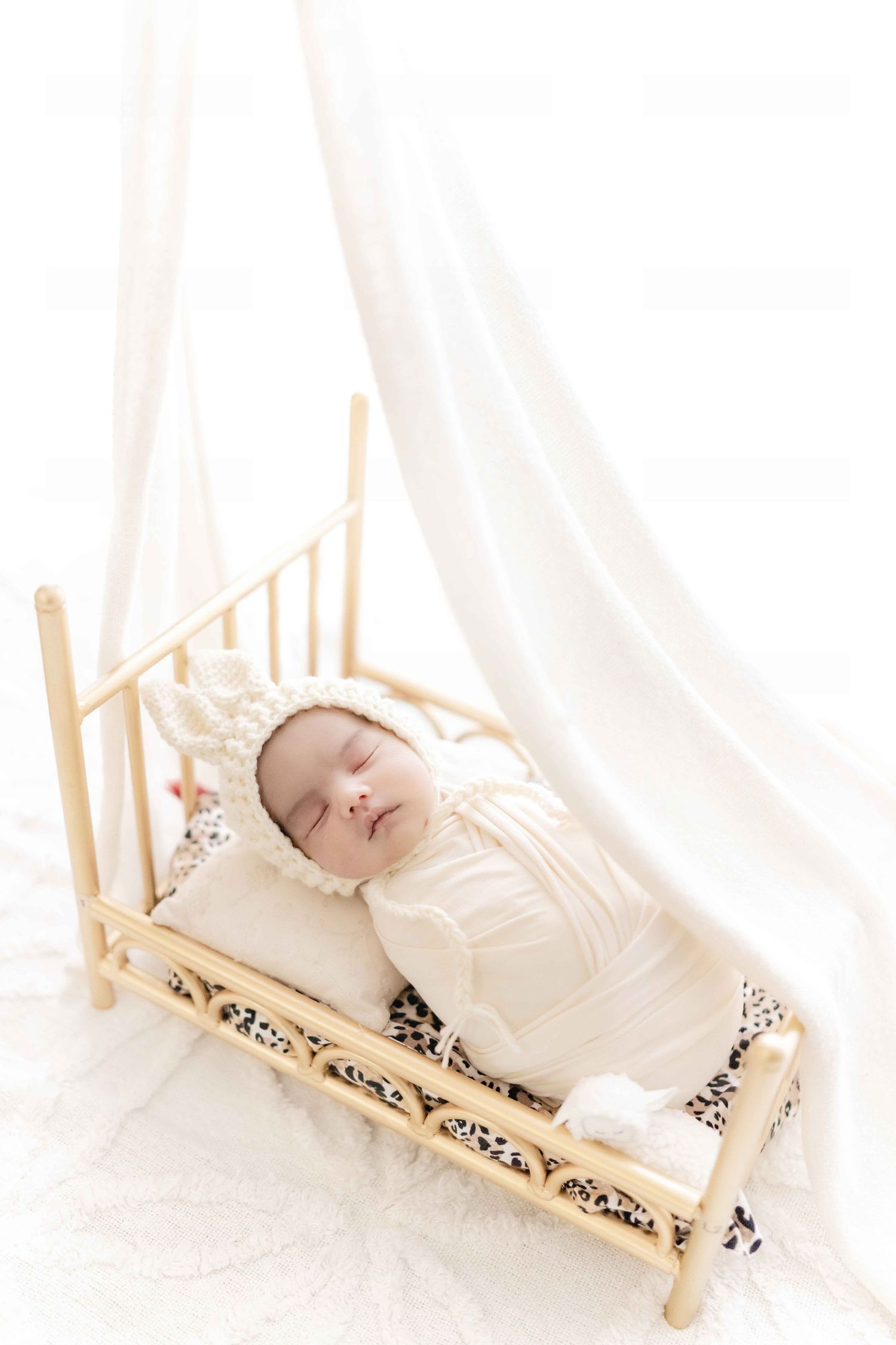 Sleeping newborn baby in cream outfit and bonnet resting in small wooden bed with white drape backdrop.