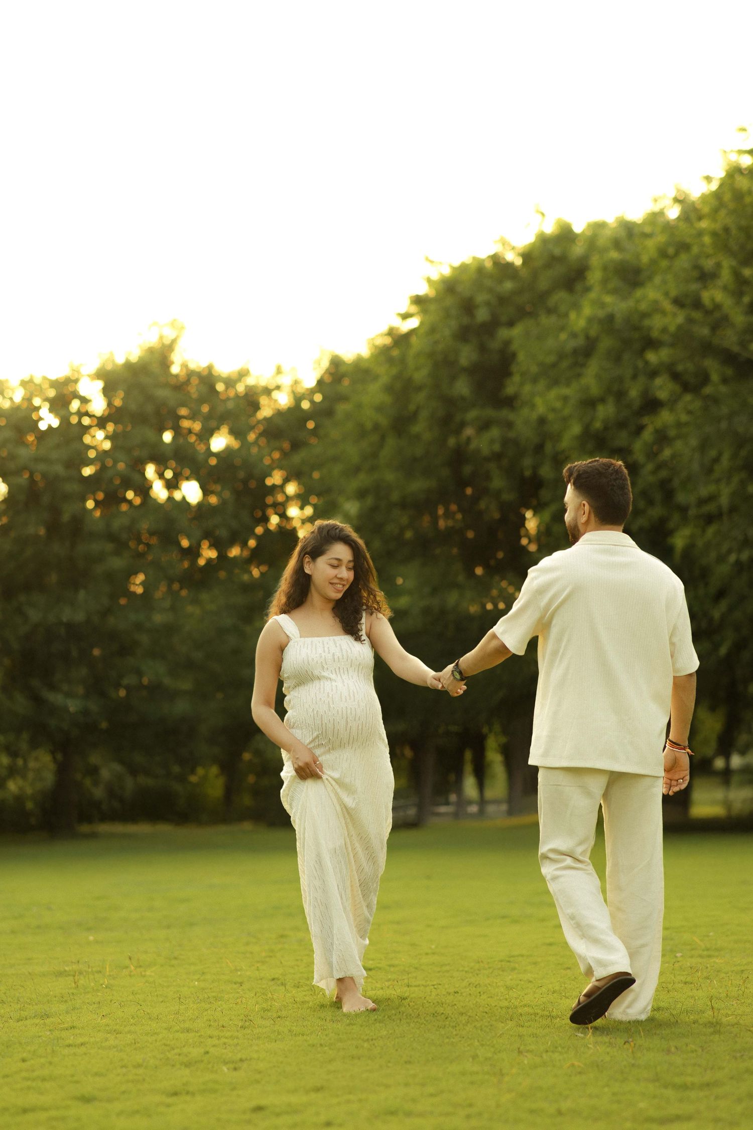 Pregnant couple holding hands in a sunlit park, wearing matching white outfits, sharing a tender moment on green grass.
