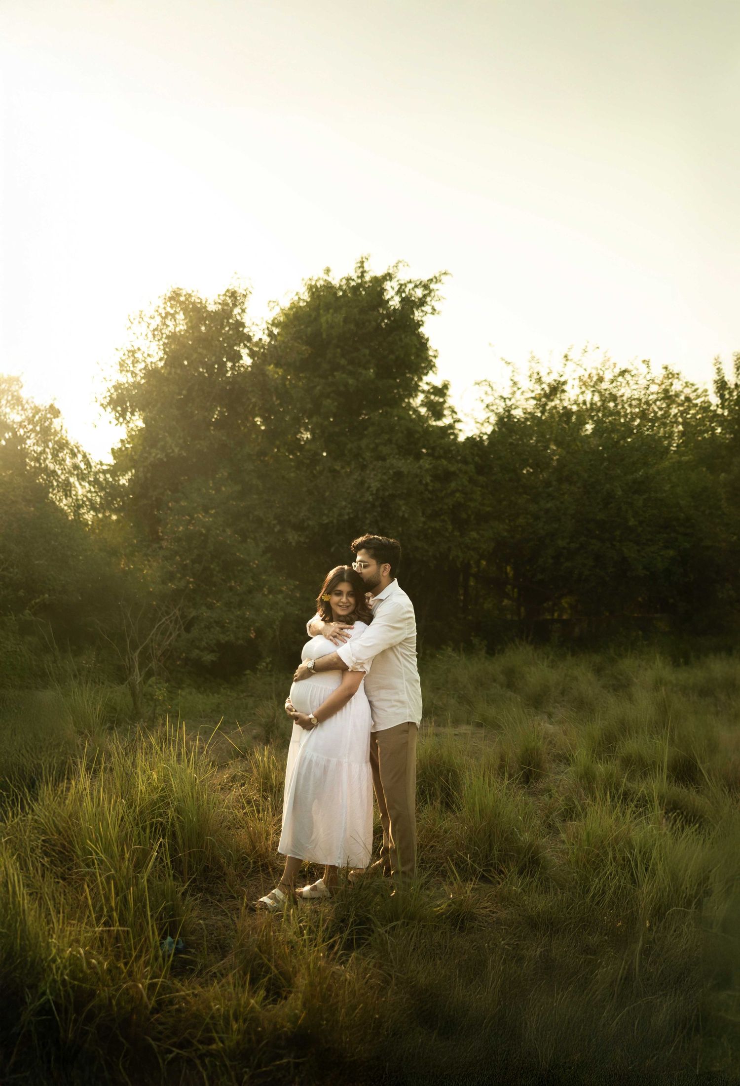 Couple embracing in golden hour light in a lush green meadow, maternity photo shoot outdoors.