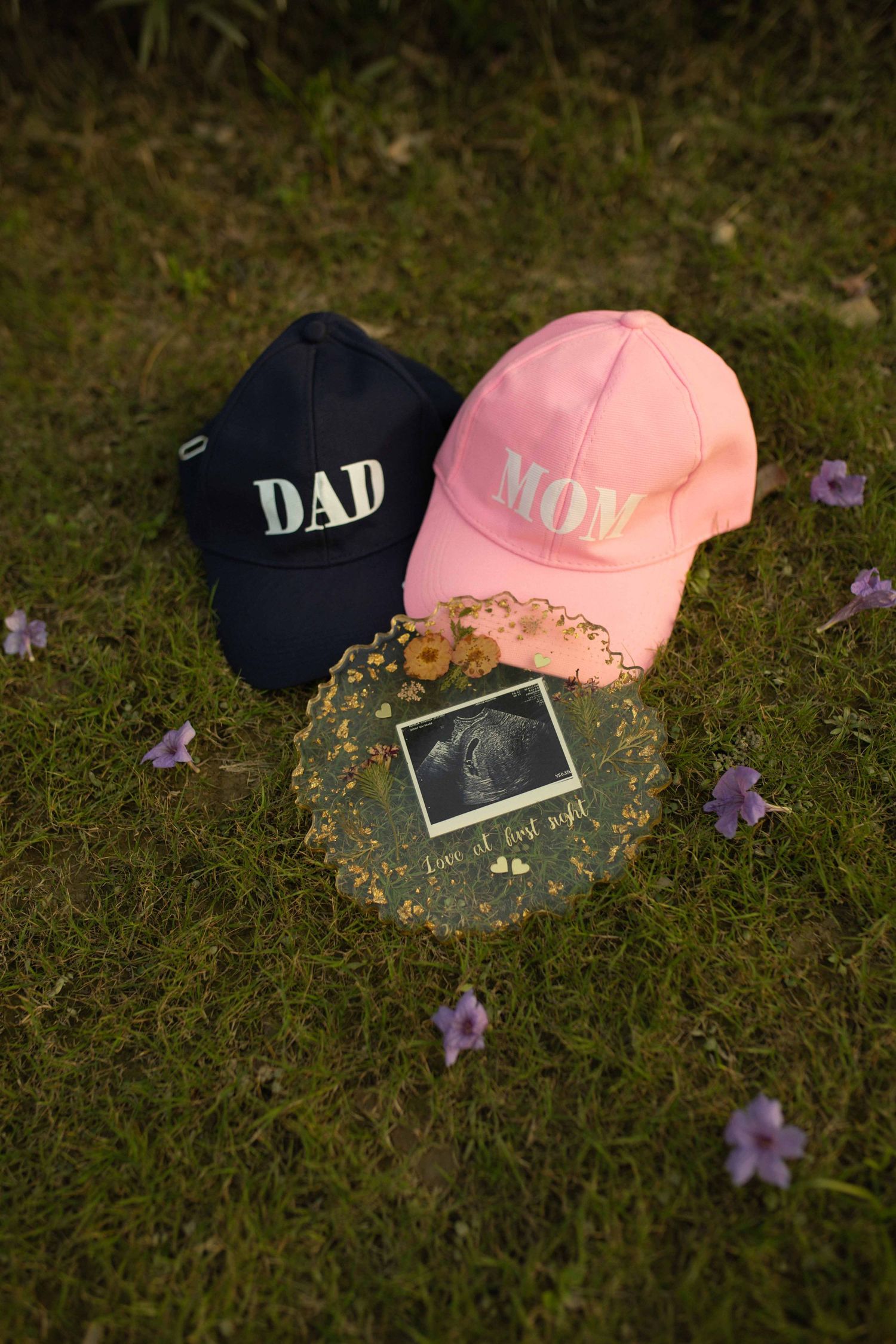 Dad and Mom baseball caps with ultrasound photo on grass, pregnancy announcement with purple flowers.